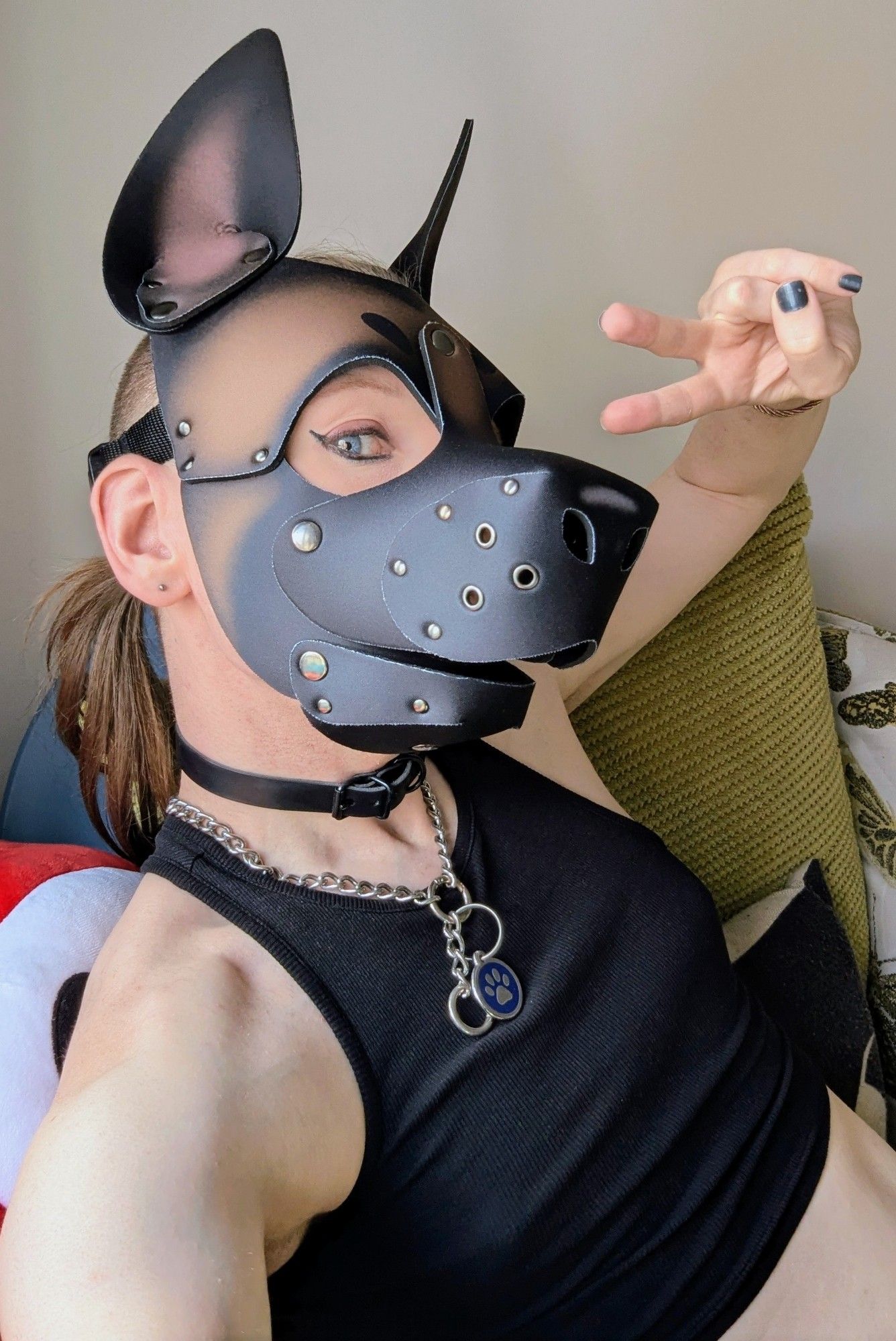 Non-binary Trans-fem sitting on a green sofa. Wearing a brown and black puppy play hood, dog chain, black cropped tank top and making a piece sign.