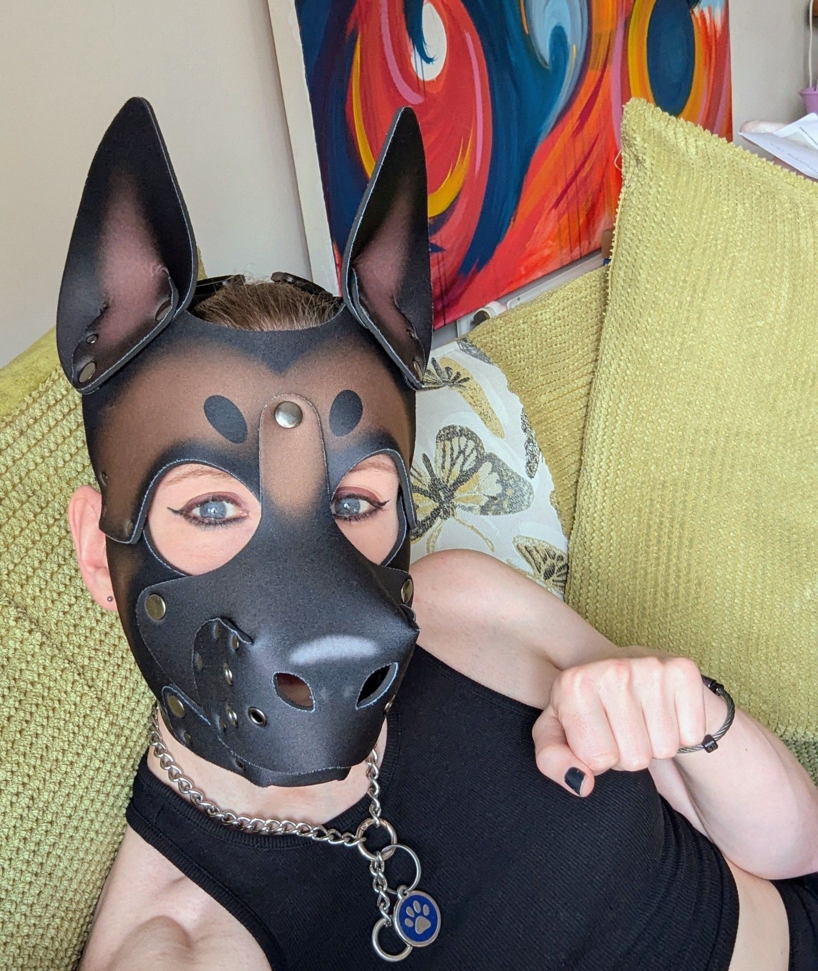 Non-binary Trans-fem sitting on a green sofa. Wearing a brown and black puppy play hood, dog chain, black cropped tank top and showing a paw.