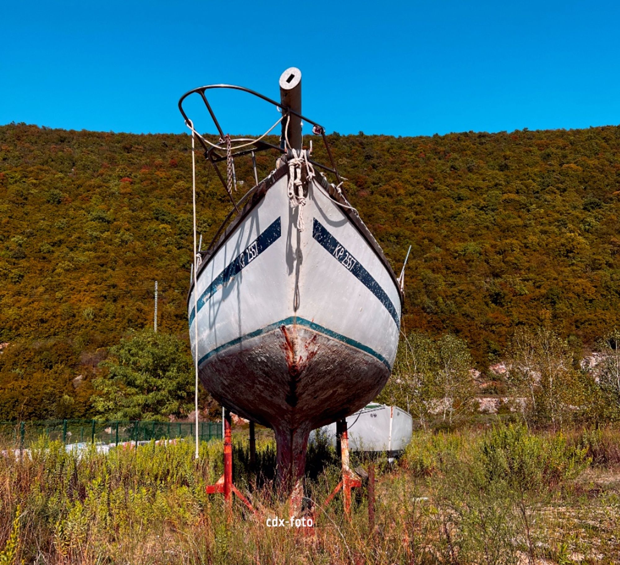 ALT-TEXT: Farbbild, Titel: Beim Spaziergang. Zu sehen ist: Ein verwahrlostes, verlassenes Boot in einem Feld am Straßenrand.