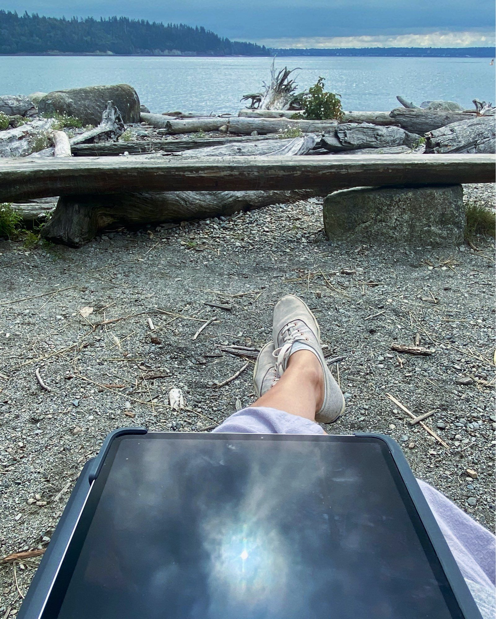 A photo from the pov of myself sitting on a bench looking out across a beach towards a blue-grey sea and sky. On my lap in the foreground rests an ipad, but it doesn’t have a normal lit screen. Instead it reflects the sun, emerging fm a hole in dark clouds, surrounded by magic looking mists and a faint ring of rainbows.