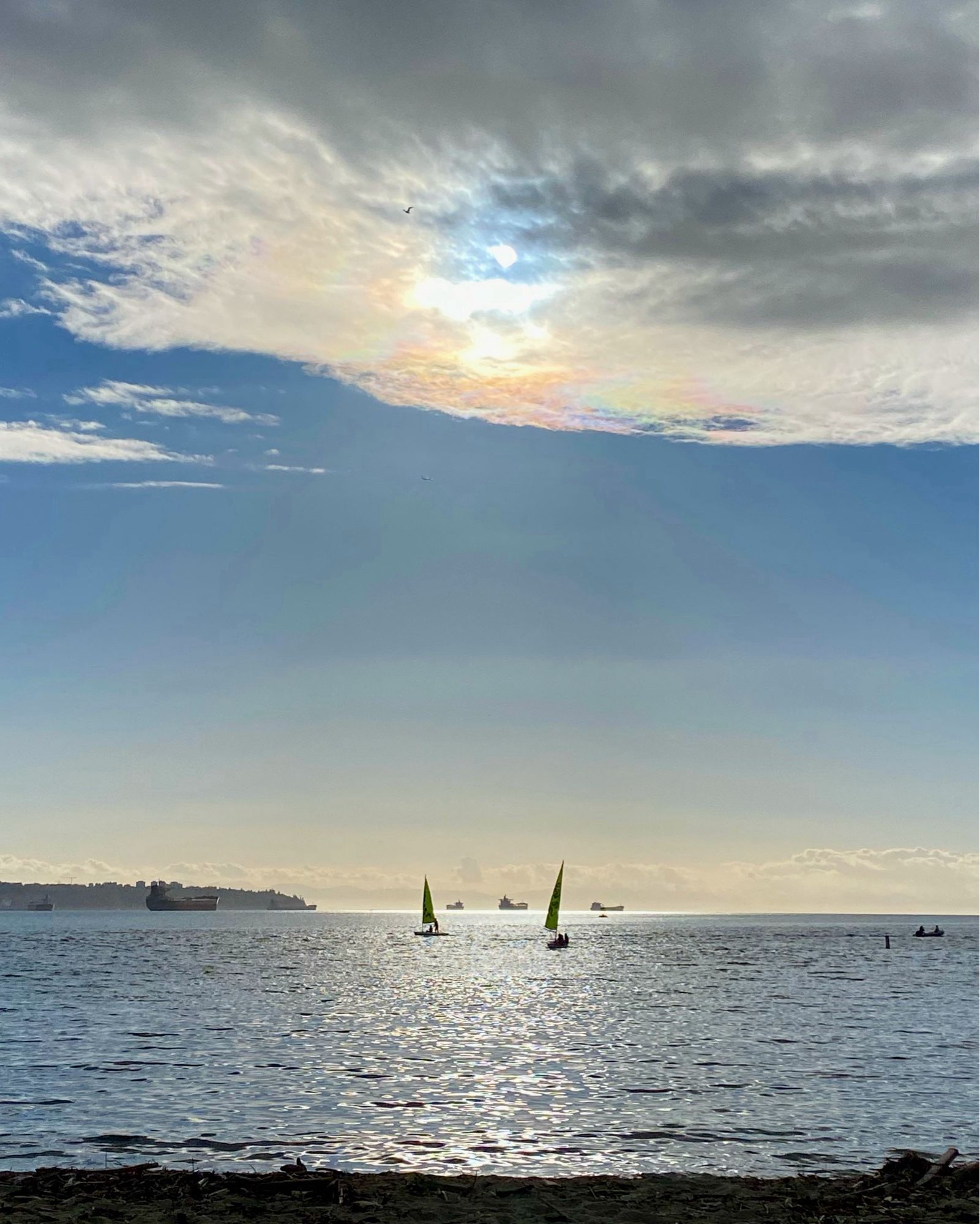 A view towards the Salish Sea, with the sun shining a silver band along anthracite waters, backlighting silhouettes of kids learning to sail and cargo ships out at sea. The sun hovers in a hole in the clouds above the ocean with an iridescent glow below it.