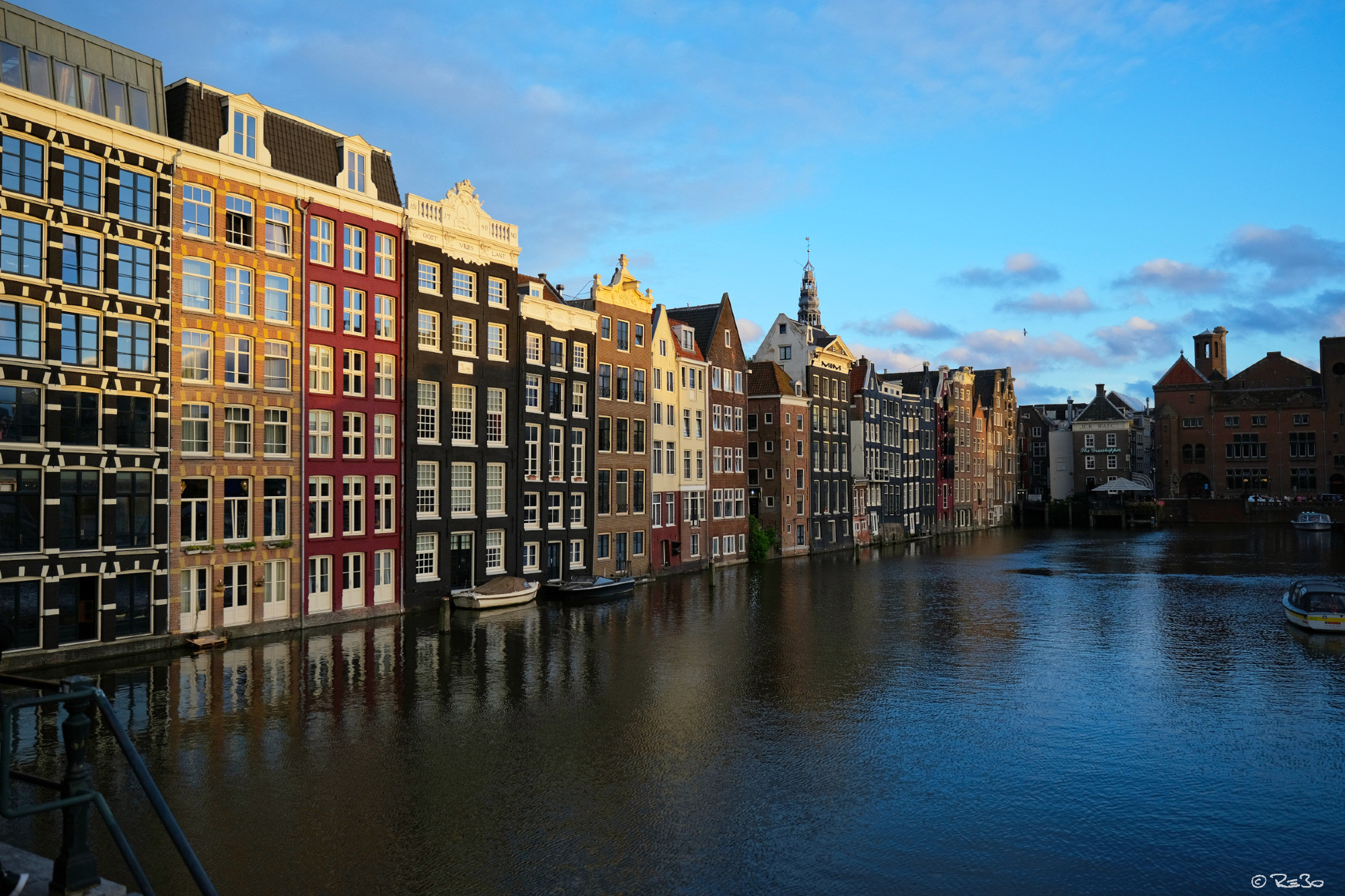 Photo of a colorful row of houses in Amsterdam's city center at the harbor basin near the Damrak. The blue water reflects the facades of the townhouses built directly on the water under a blue sky.