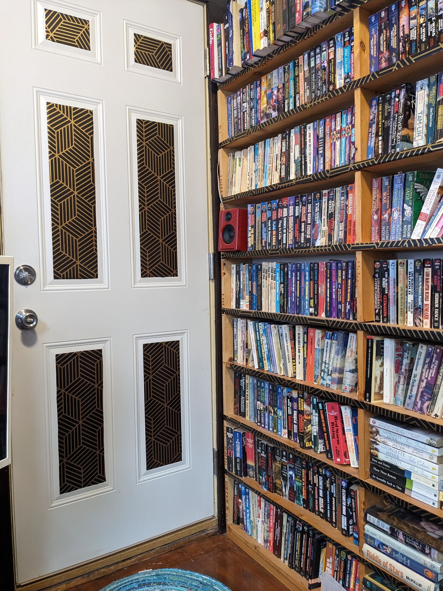 A white exterior door in the 6-panel style has had black and gold geometric paper applied to the inner panels. If you look closely, the front edges of the wooden bookcase are also covered with the paper .