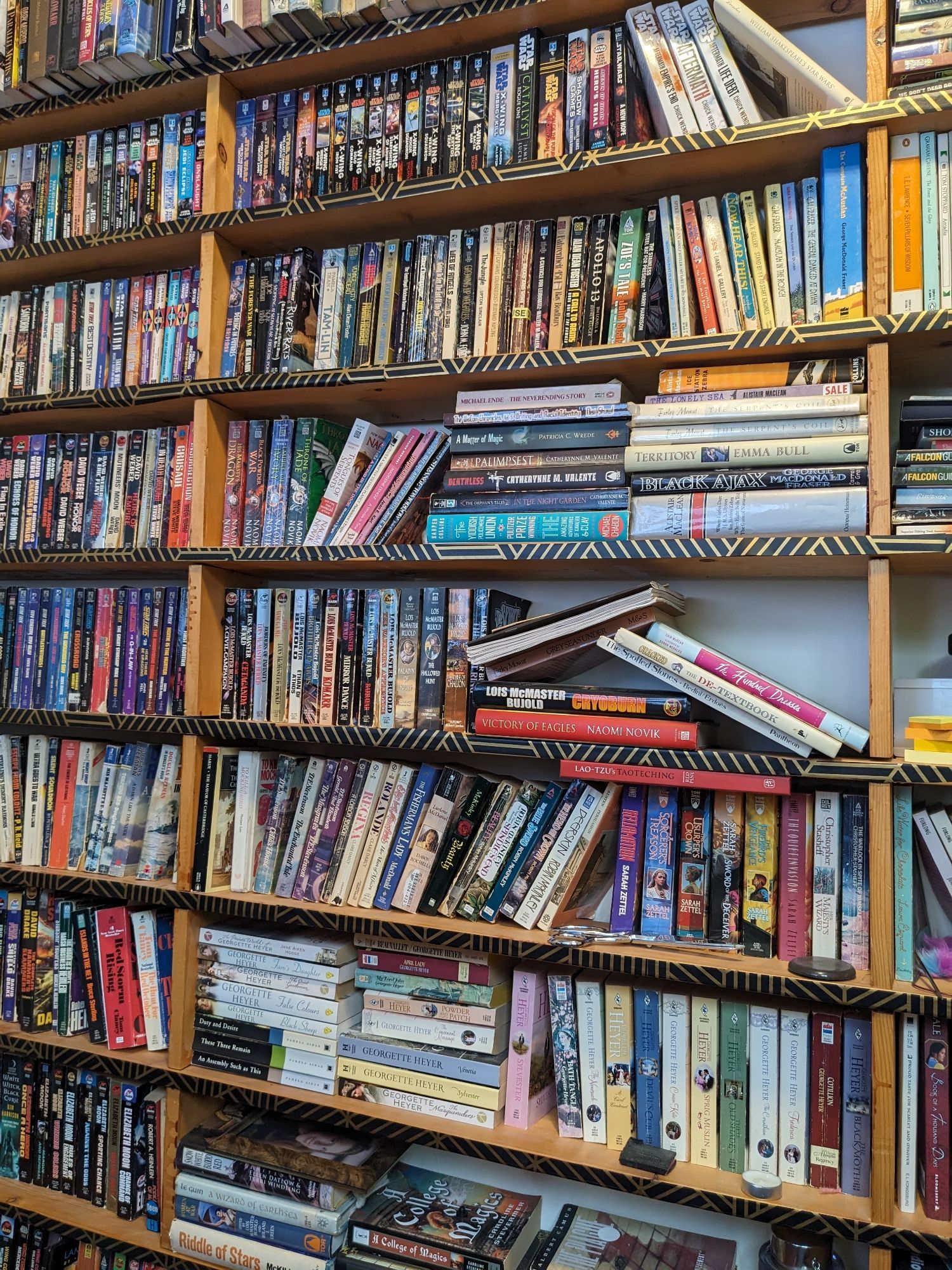 A bookcase full of all sorts of books, including Heyer, Bujold, Novik, and a huge number of Star Trek and Star Wars books. The horizontal edges of the bookcase have been trimmed with black and gold paper that mostly shows gold lines.