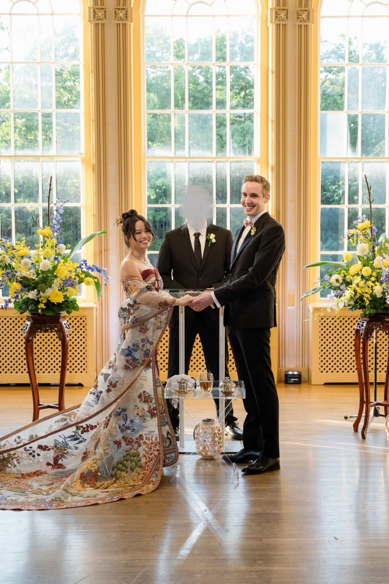 A couple in front of an officiant flanked by flowers, bride is wearing a CUSTOM cheongsam-style wedding gown