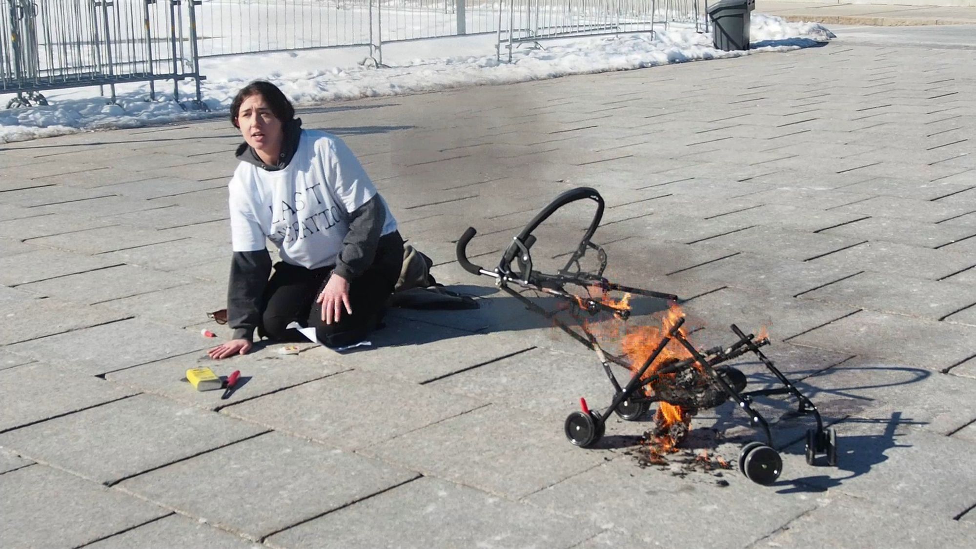 A young woman in a white "Last Generation" t-shirt sits on the pavement outside Parliament with her hand glued to the ground while a baby stroller burns next to her.