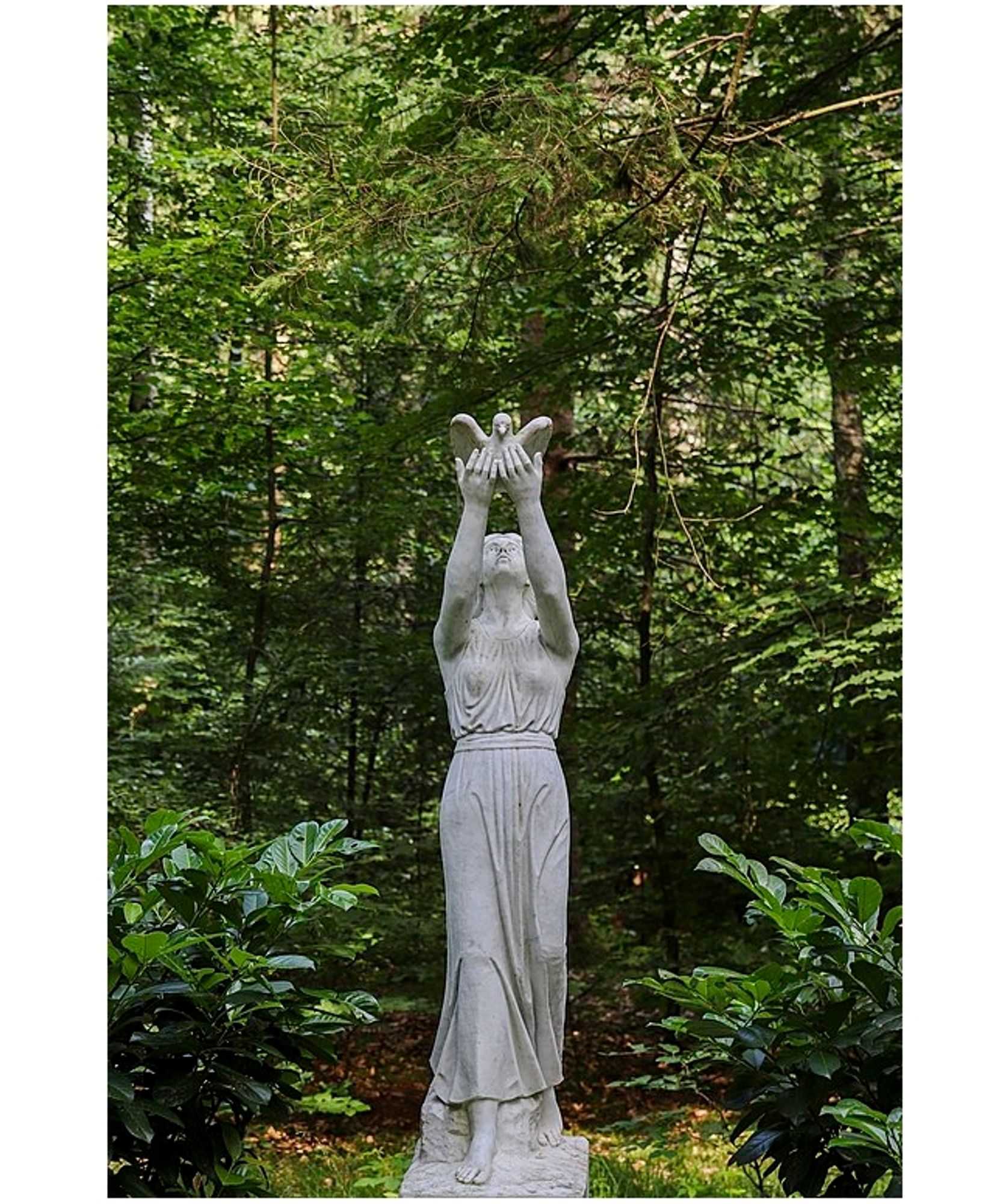 Photo in woodland of a grey stone statue in the form of a woman in a blouse and long skirt with her arms raised high releasing a dove. The photo is titled "rebirth and hope".