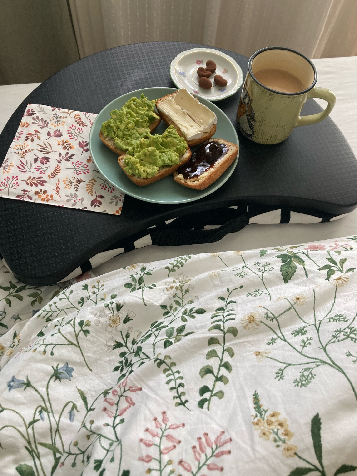 Knee tray in bed. Bun halves with avocado, camembert cheese and dark jam. Small plate with cocoa-powdered nuts. Mug with tea.