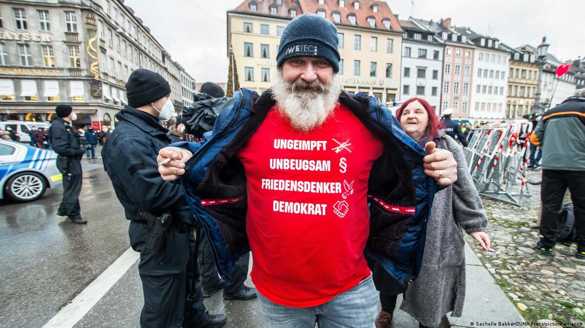 Ein Demonstrant dumm. Mann mit Bart und schwarzer Wollmütze am Rande einer Querdenkerdemo. Er öffnet seine Jacke und hält sie so, dass man die Aufschrift seines Shirts lesen kann. Darauf steht Ungeimpft, unbeugsam, Friedensdenker, Demokrat.