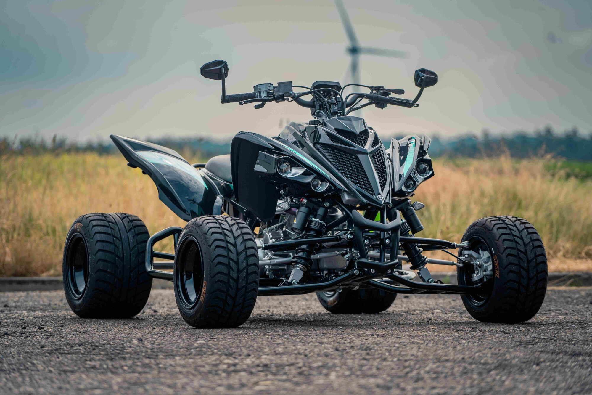 Ein Quad fotografiert auf einer geteerten Straße vor einem Feldweg. Im Hintergrund unscharf ein Windrad.