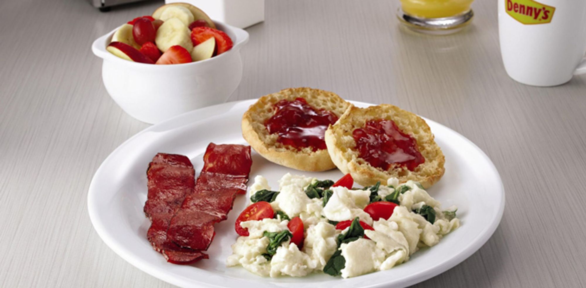 2 strips of canadian bacon with scrambled egg whites, tomatoes and spinach, next to a toasted English muffin with red jam, and a bowl of apples, strawberries, and banana slices,