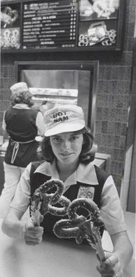 A woman in a Hot Sam uniform holds up two pretzels with tongs, one in each hand