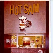 At a mall food court stand, a woman prepares pretzels over a brick facade. Above her is a sign with a little man in white, holding up a large pretzel