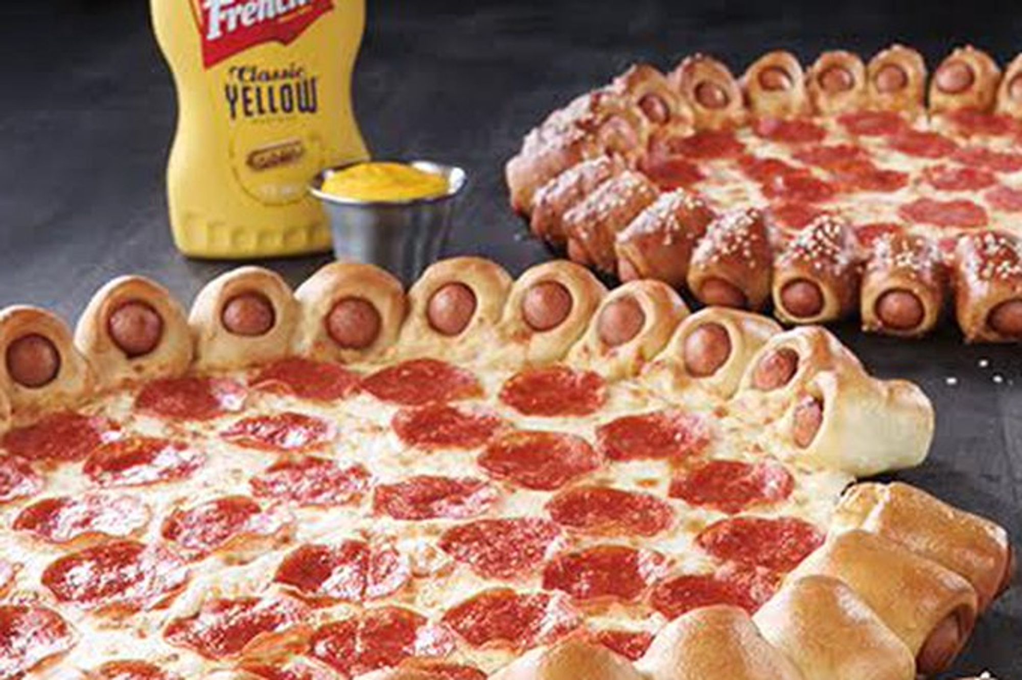 A pepperoni pizza, with mini hot dogs baked into the crust all around the edge. a bottle and small metal cup of French's yellow mustard are in the background