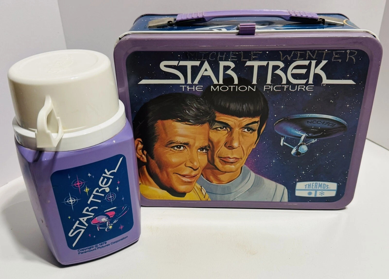 A metal purple lunchbox with an illustration of Kirk and Spock, with outer space and The Enterprise in the background, below the words "Star Trek The Motion Picture". Next to the lunchbox is a purple plastic thermos with white top, that says "Star Trek" across an outer space scene