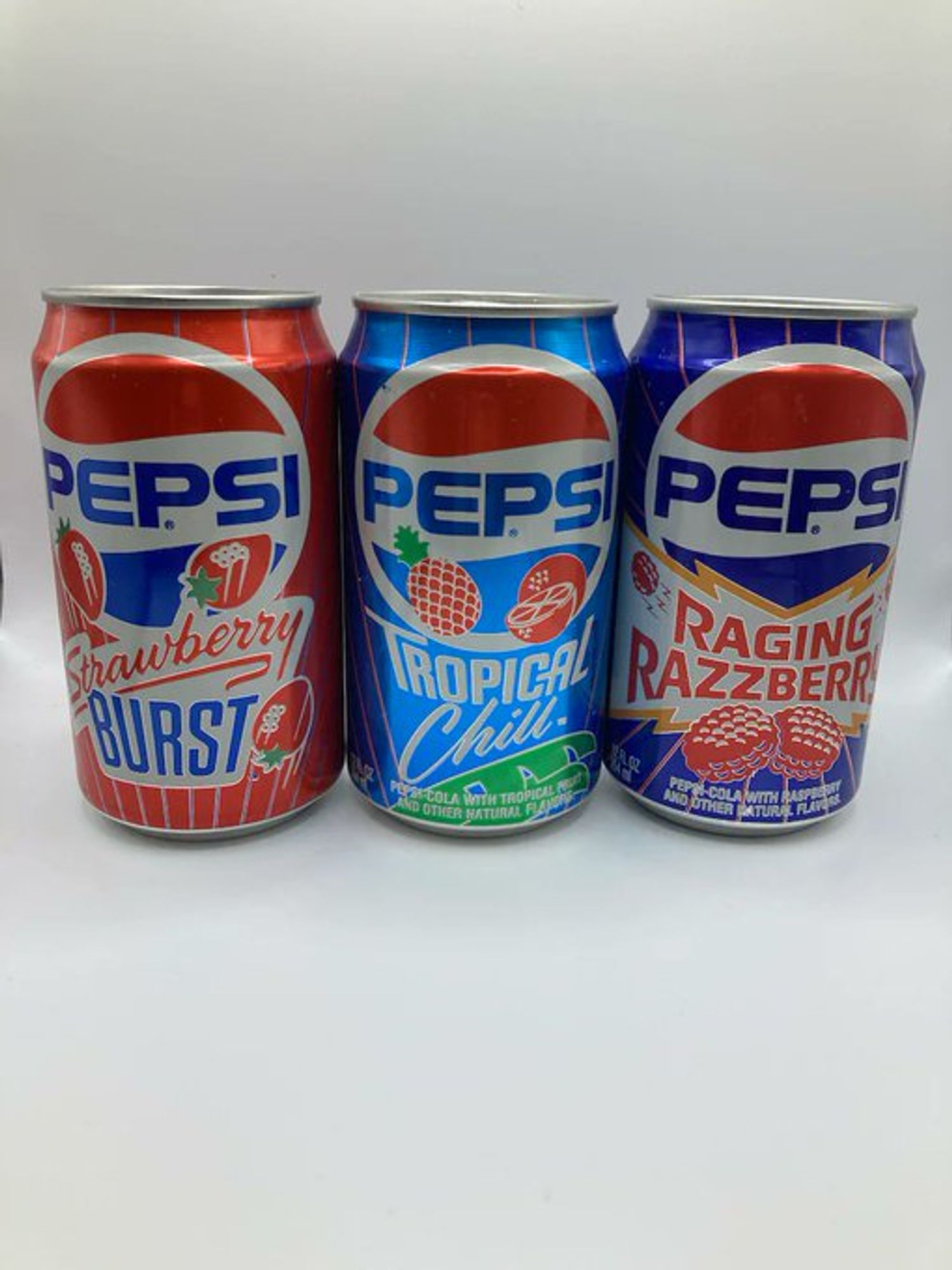 A red Pepsi can with three strawberries flying around on it, a blue Pepsi can with a pineapple and halved orange, and a dark blue Pepsi can with two large raspberries on it