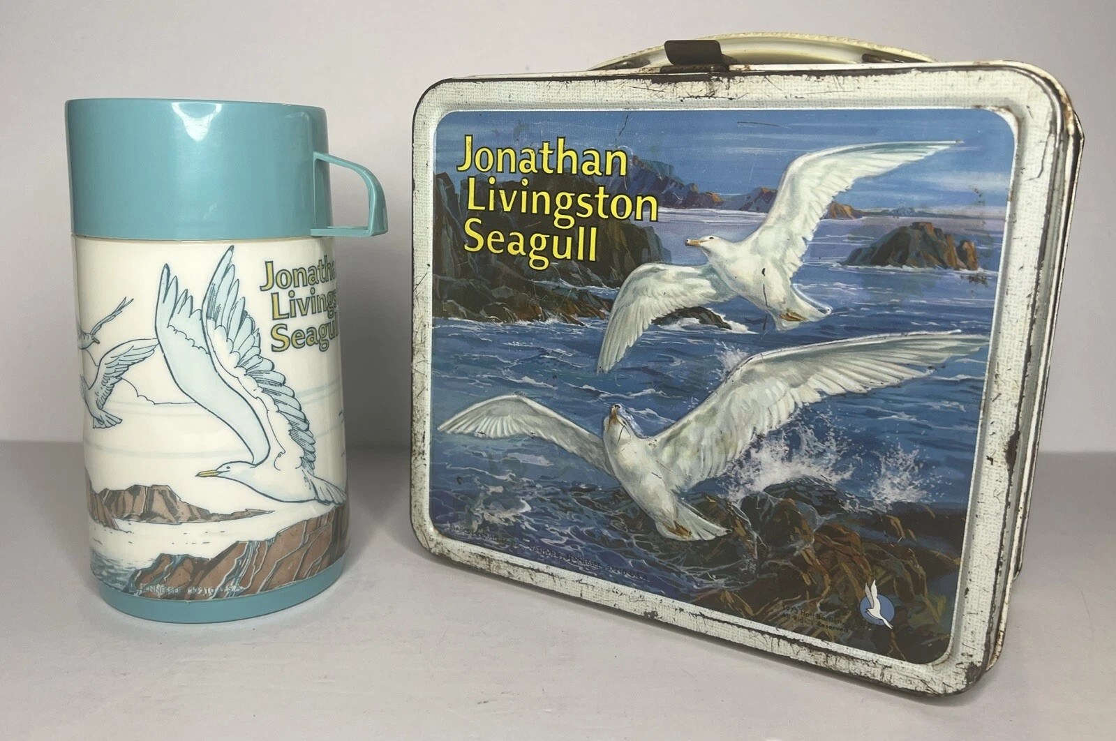 A white metal lunchbox with a lovingly illustrated image of 2 seagulls flying over a rocky inlet as waves crash. Next to it is a white thermos with a teal lid, and illustrations of flying seagulls next to the words "Jonathan Livingston Seagull"