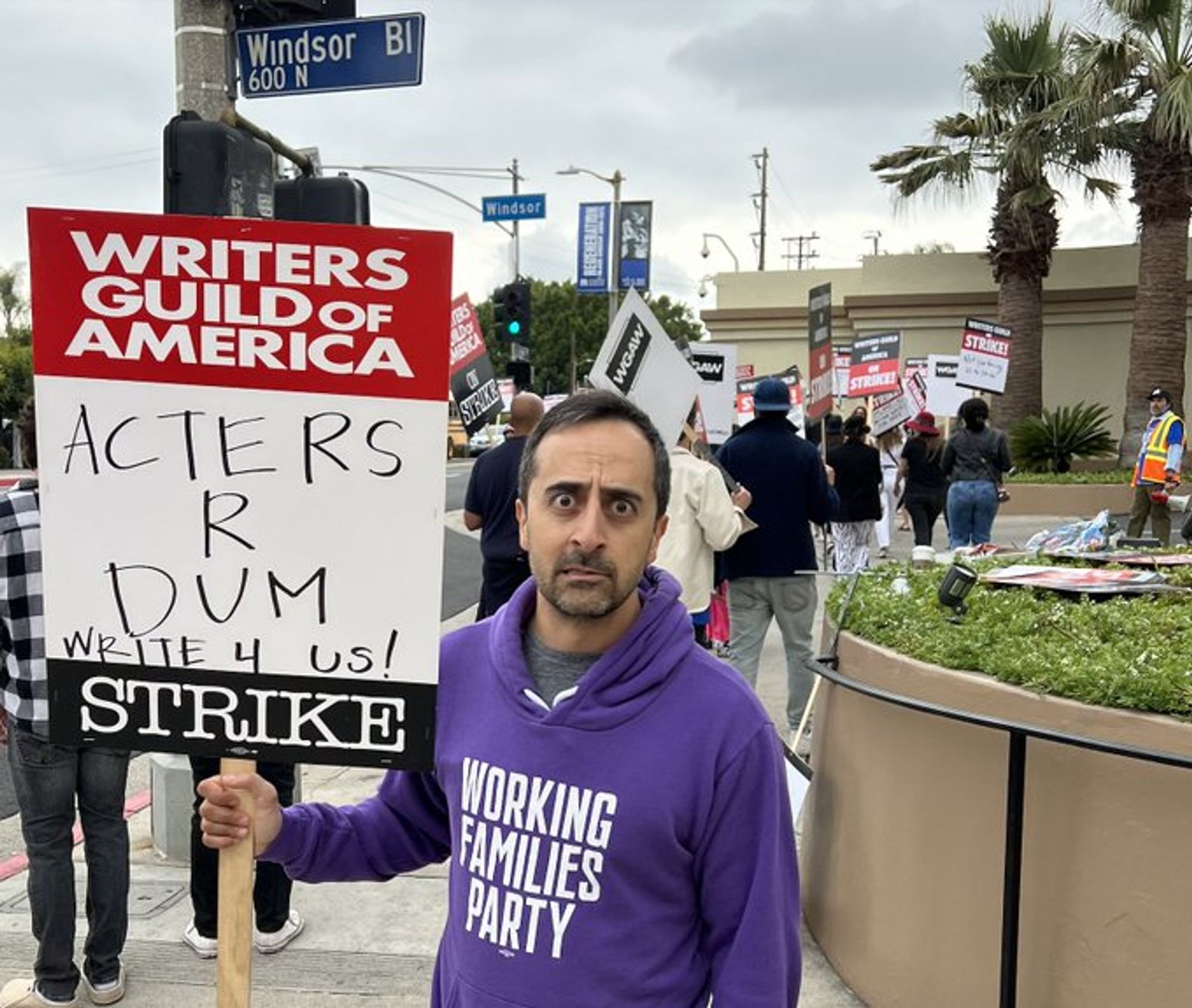 Person (Amir Talai) wearing a Working Families Party sweatshirt and carrying a sign for the Writer's Guild that says Actors r dum write 4 us