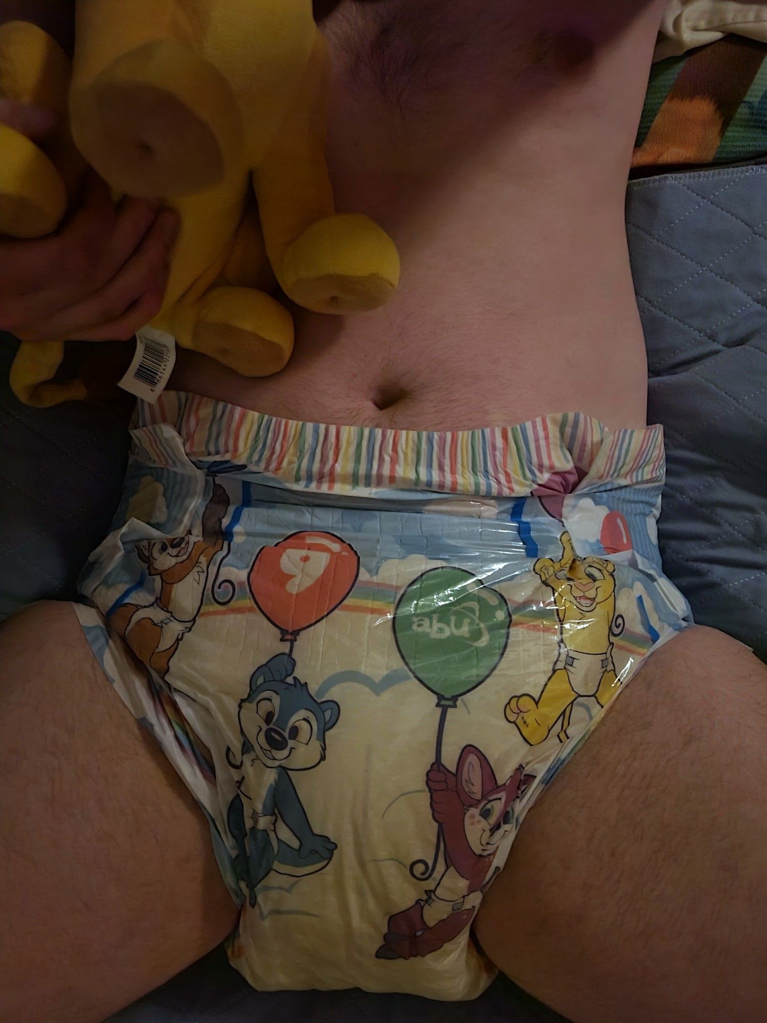 A dimly-lit kitty all cozy in bed with his Simba plush and very droopy Tiny Tails diaper. The tapes are straining a bit, such that if an adult were present, changies would be likely.