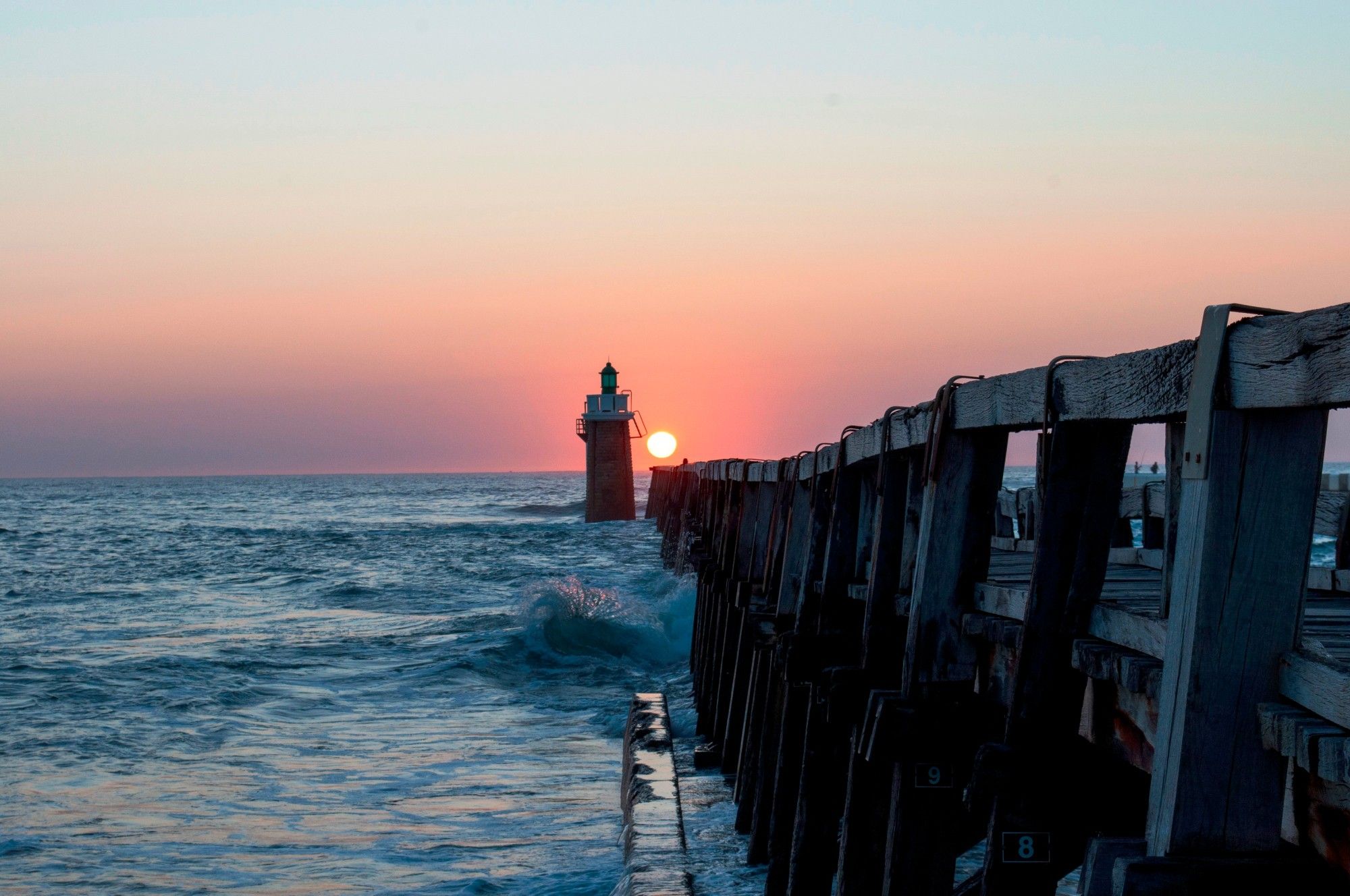 une estacade en bois au bord de l'ocean avec un phare, le soleil couchant juste à son extrémité