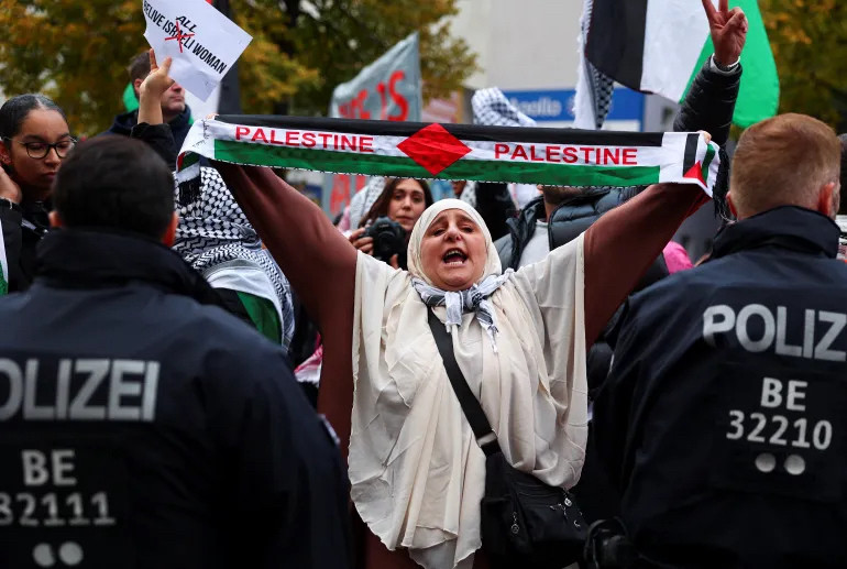 Police loom heavily in foreground as a protestor wills their stance larger, bigger, and wider to overcome the oppressive presence with arms thrust wide to hold taut a banner for Palestine that says “Palestine, Palestine” as if crying out, wailing and weeping, to bolster the inherent and uncontainable power of their stance.