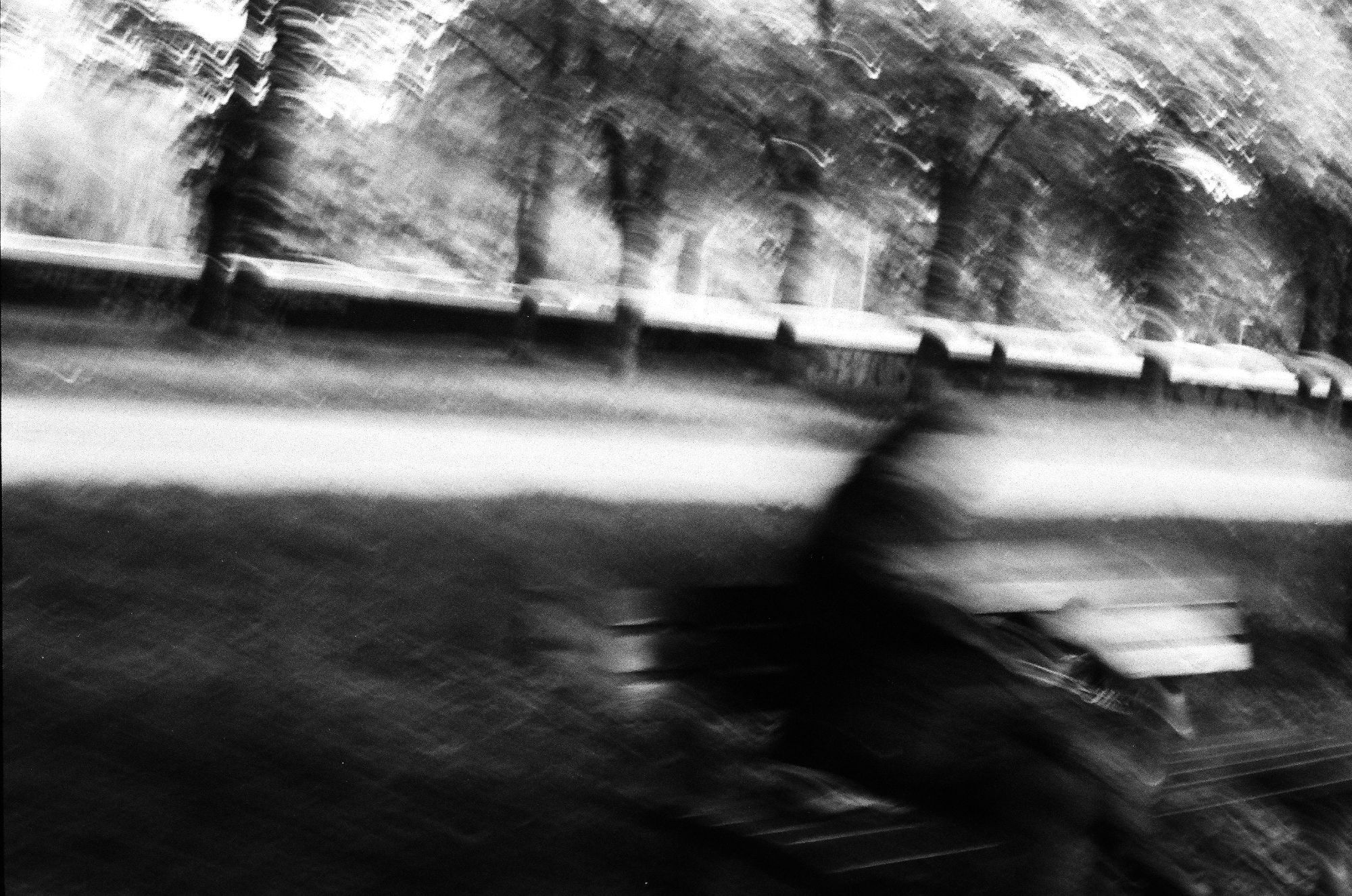 a passing by, someone you might never interact with or come across again, yet still lingering in a corner of your mind

a blurry black and white photo of a person sitting on a bench, face blurred out, a path and alley of trees in the background