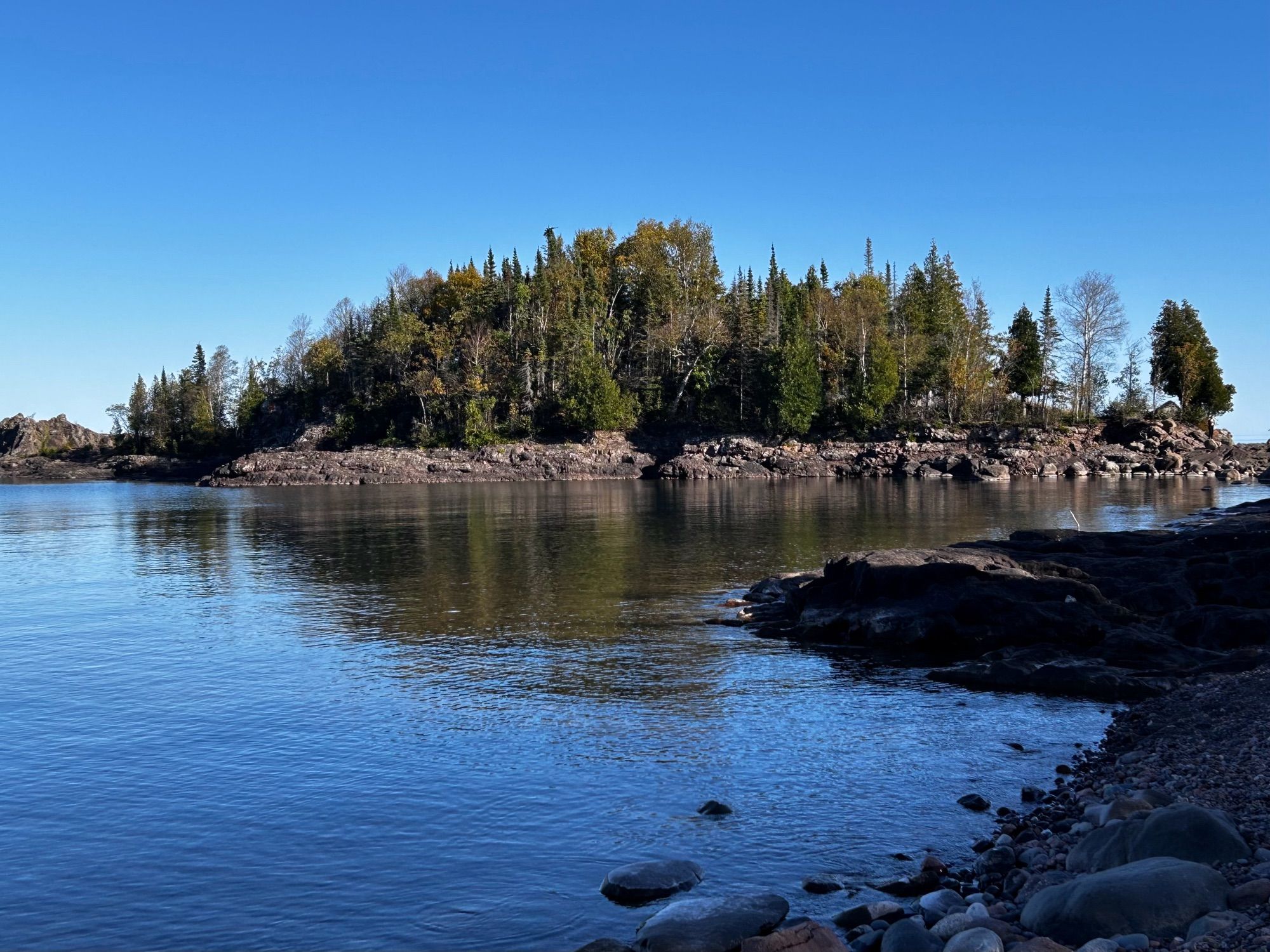 View across calm blue water of a small tree covered isthmus with blue sky above.