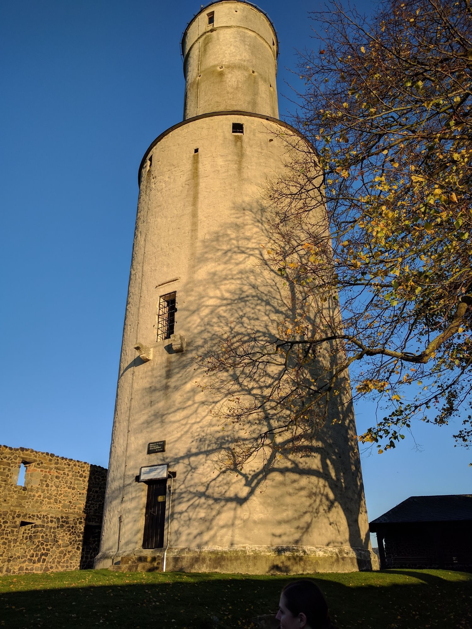 Grauer ,schlichter Turm, der oben drauf noch einen kleineren Turm hat. Man sagt, er habe eine Butterfass-Form. Blauer Himmel. Am rechten Bildrand ein Baum mit nur noch wenigen gelben Blättern. Der Schatten des Baumes ist auf dem Turm zu sehen. Links ein Stück Burgmauer