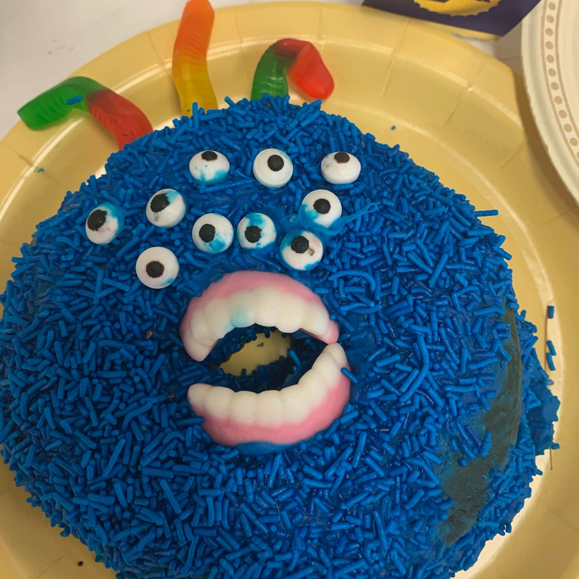 Photo of a homemade confection from a county fair. It appears to be a large donut covered in blue candy sprinkles, with 10 candy eyeballs and a set of candy dentures arranged as though screaming.