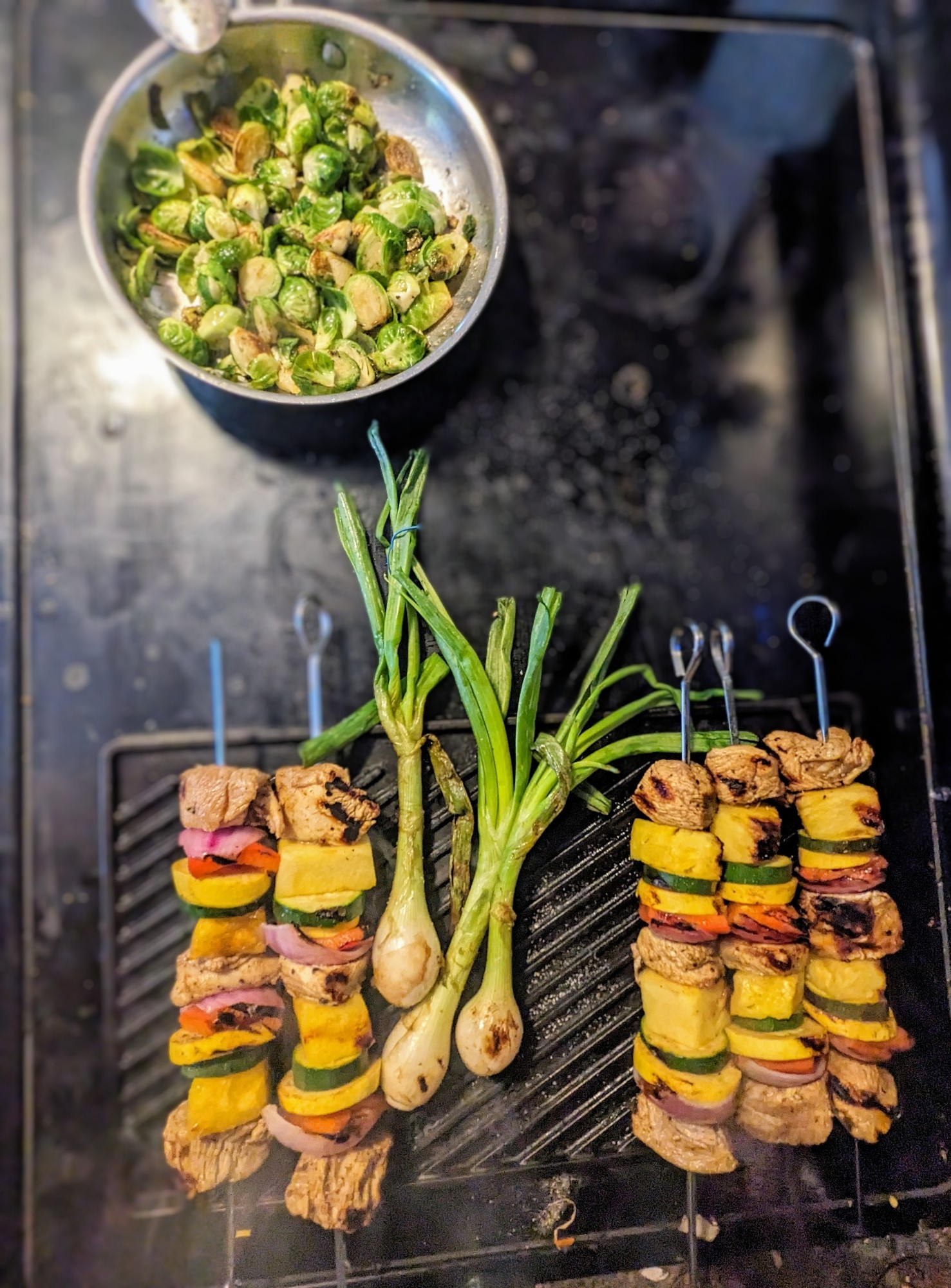 A skillet of Brussels sprouts and a cast iron griddle with 3 cebollitas and 5 chicken and veggie kebobs sitting in a dirty glass stovetop.