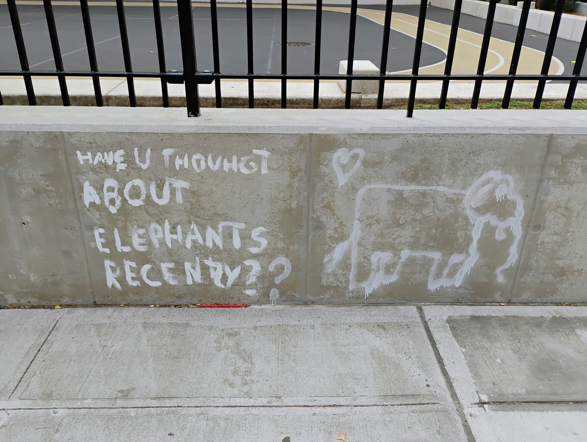 A concrete slab wall next to a sidewalk. "HAVE U THOUGHT ABOUT ELEPHANTS RECENTLY??" is written on the left, while an elephant and a heart shape is drawn on the right, both in white paint.