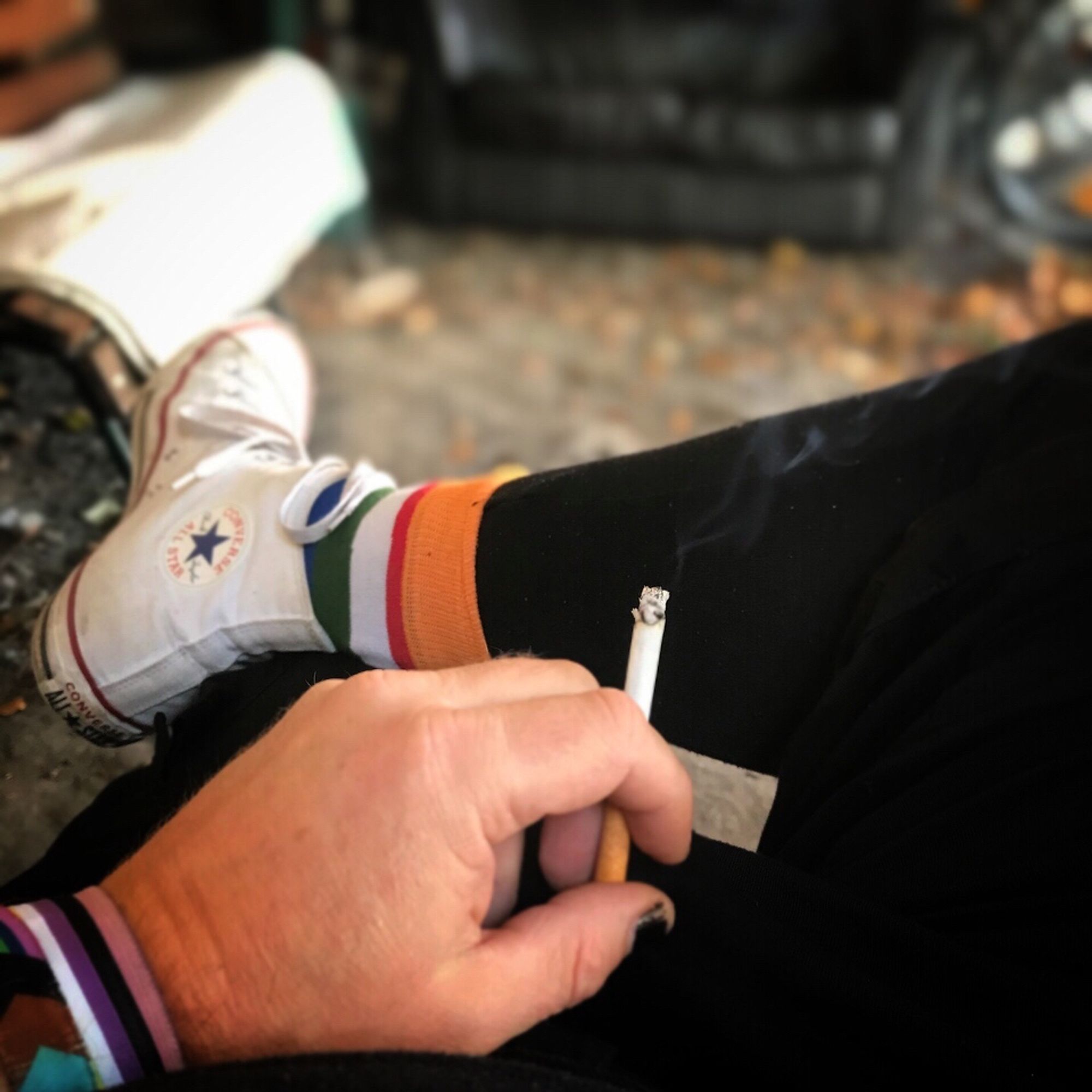 Point of view: ich sitze in der Raucher*innenecke, mein rechtes Bein überschlagen. In der linken Hand eine Zigarette. Du siehst schwarze Leggings mit schwarzer kurzer Hose, weisse chucks, Ringelstrümpfe und ein Non-binary Band am Handgelenk. Der (sichtbare) Daumennagel ist schwarz lackiert. Im verschwommen Hintergrund ein altes Sofa