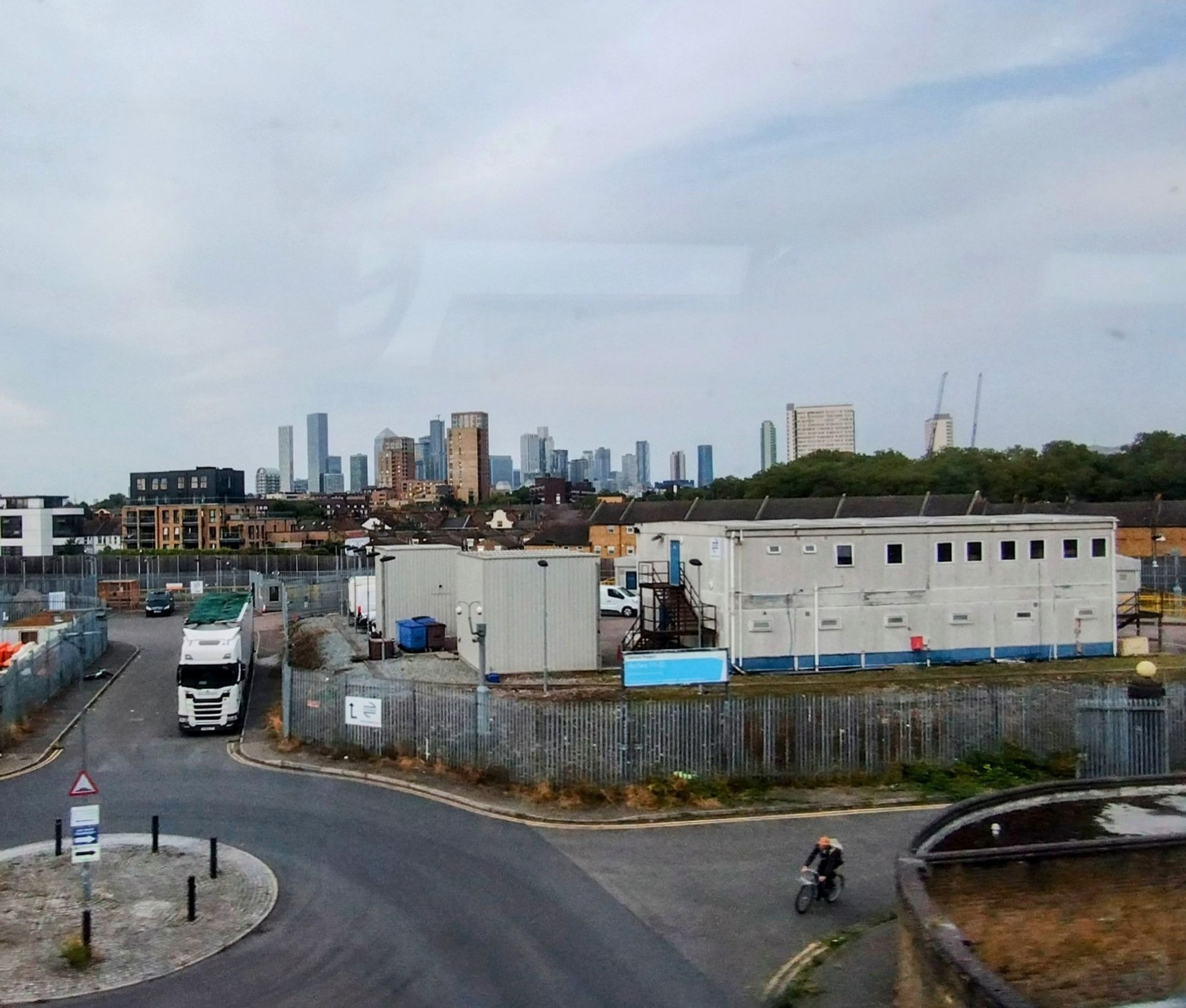 In the distance Canary Wharf sky scrapers huddle on the skyline. In the middle ground an industrial estate, prefab buildings, a maze of iron fencing. A lorry is parked up and in the foreground there's a roundabout. A cyclist is just emerging from a road on the right to go round it.