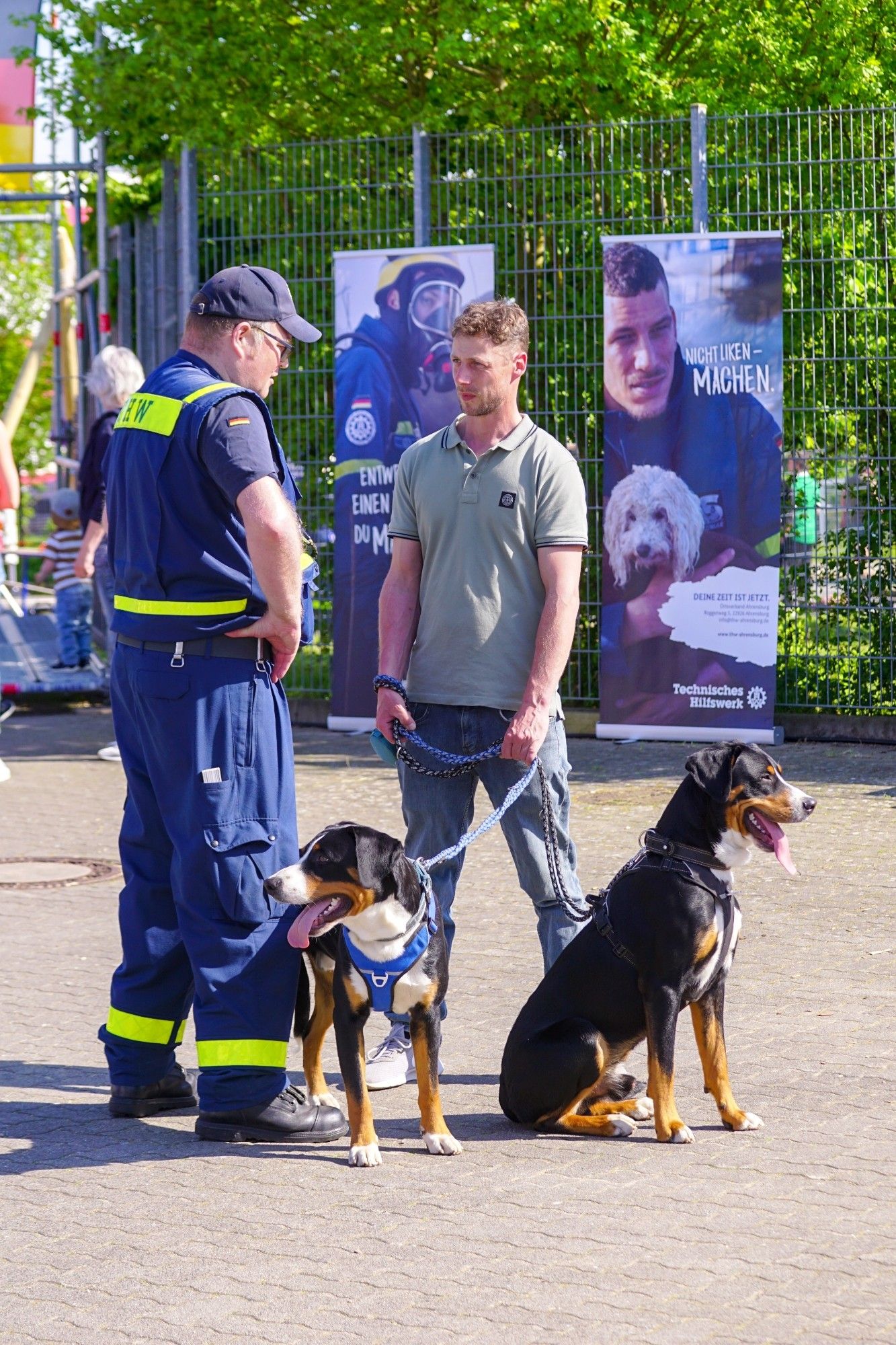 Ein Besucher spricht mit einem THW-Helfer. Der Besucher hat zwei Hunde an einer Leine dabei. Im Hintergrund ist auf einem Plakat ein THW-Helfer mit Hund abgebildet sowie der Überschrift "Nicht liken - machen."