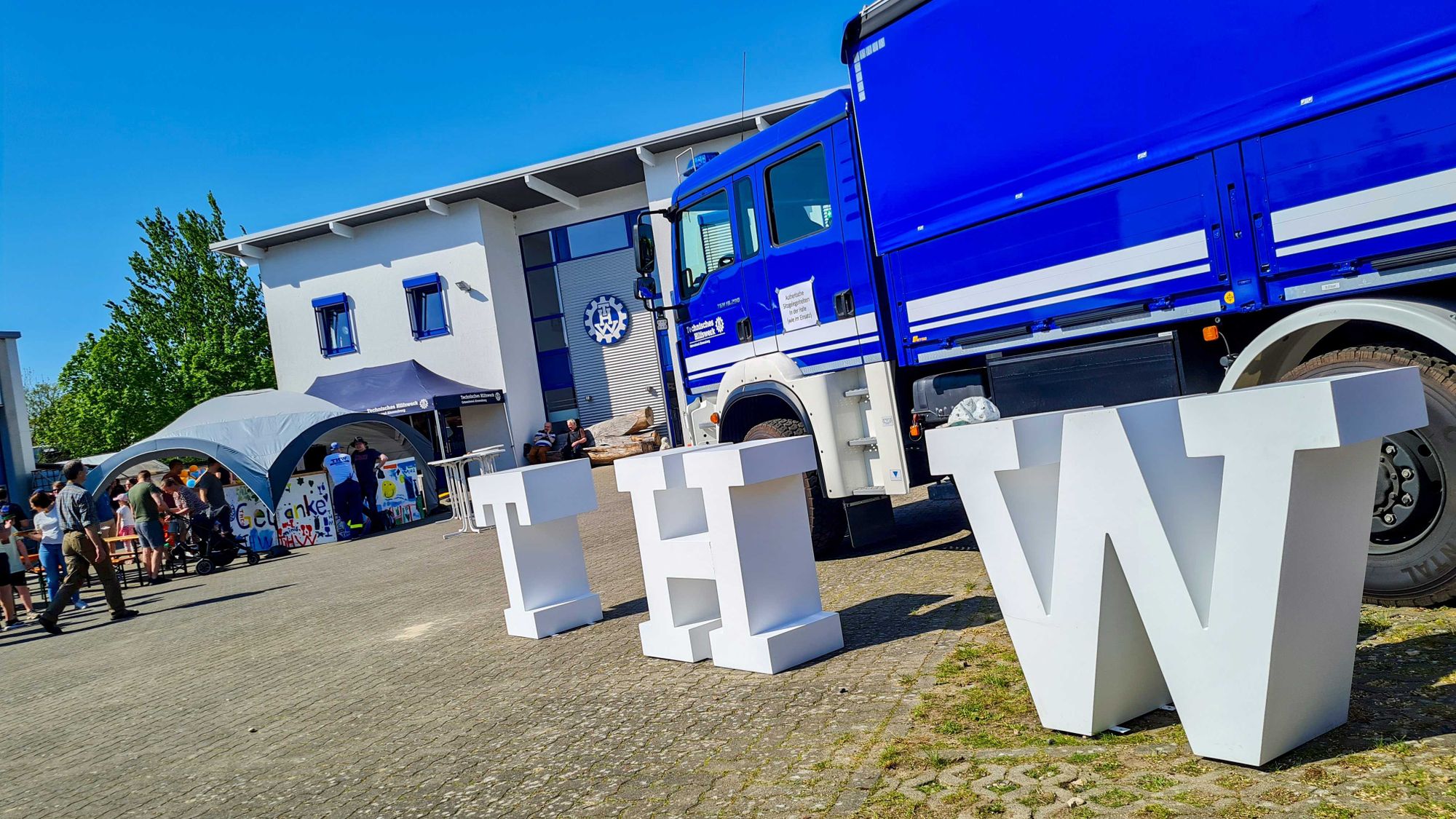 Drei brusthohe weiße Buchstaben T, H und W stehen vor einem blauen LKW vom THW. Im Hintergrund ist die Unterkunft des THW Ahrensburg zu sehen sowie links ein Getränkestand.