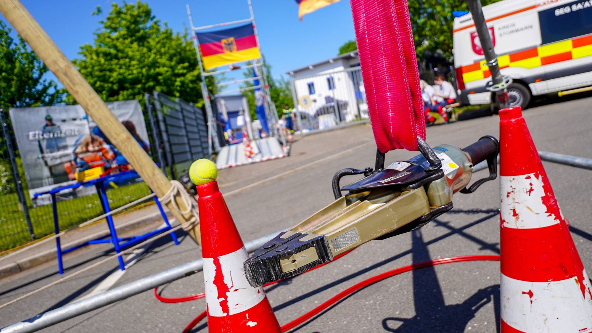 Ein hydraulischer Greifer hängt in einem sogenannten Dreibock. Auf einem Absperrkegel daneben liegt ein Tennisball. Im Hintergrund ist unscharf die Hofeinfahrt zum THW-Gelände zu sehen.