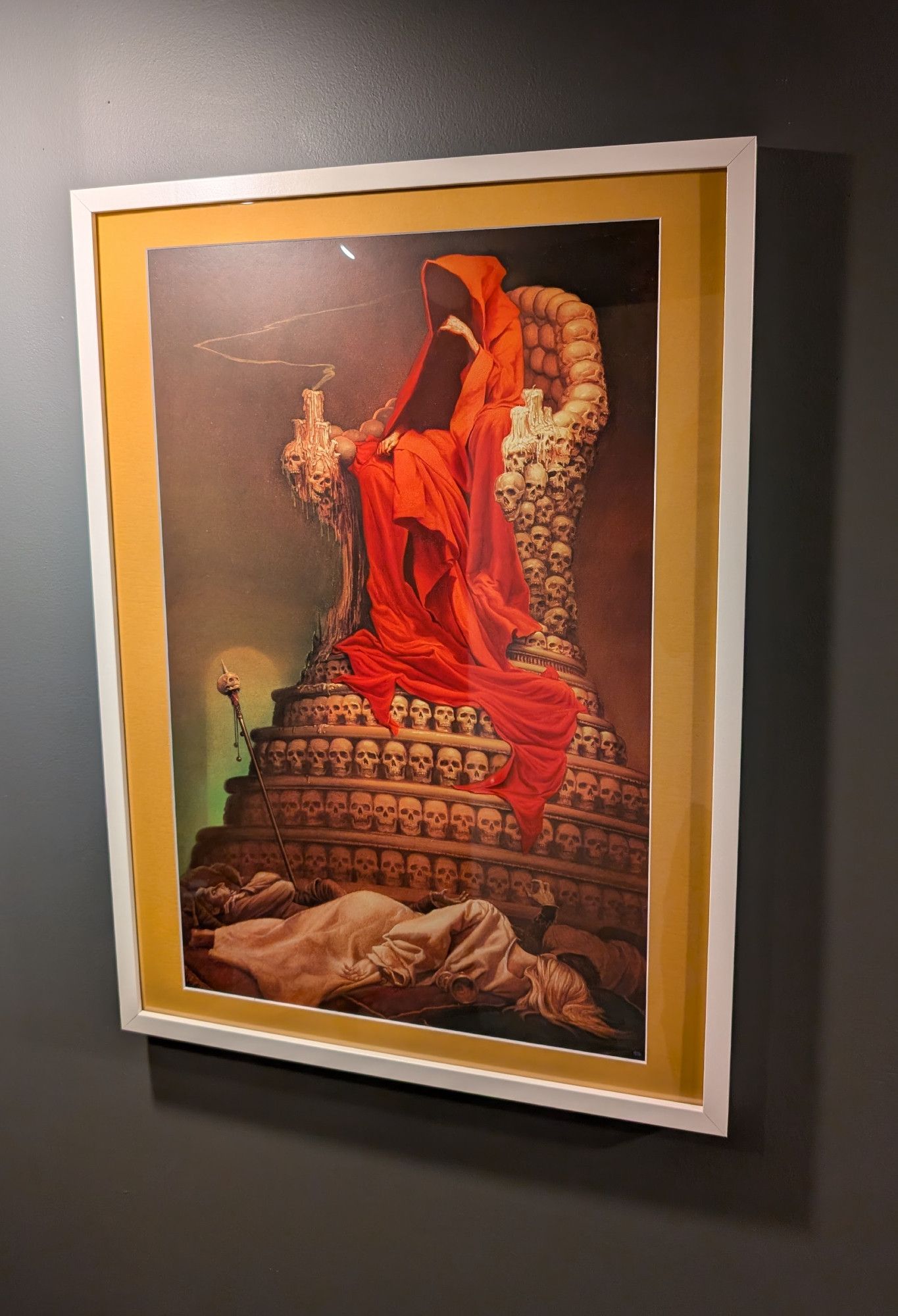 Michael Whelan's "THE CRIMSON KING" hung and framed on a wall with an off-yellow border. The work depicts a figure robed in red sitting atop a throne comprised of skulls overlooking several corpses.