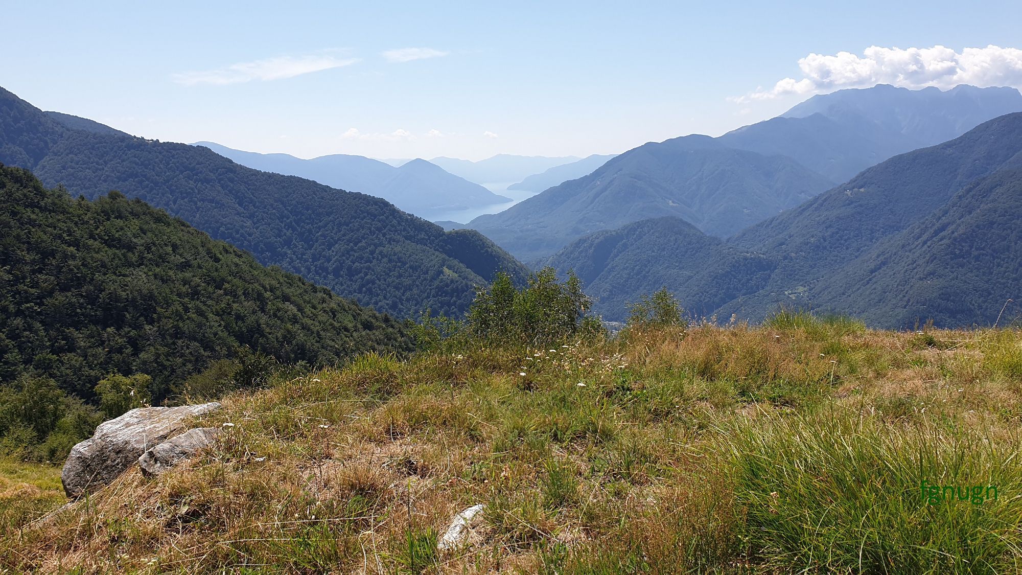 Ausblick von einer Wiese in den Bergen über das Maggiatal. Im Hintergrund ist der Lago Maggiore zu erkennen. Die Sonne scheint.