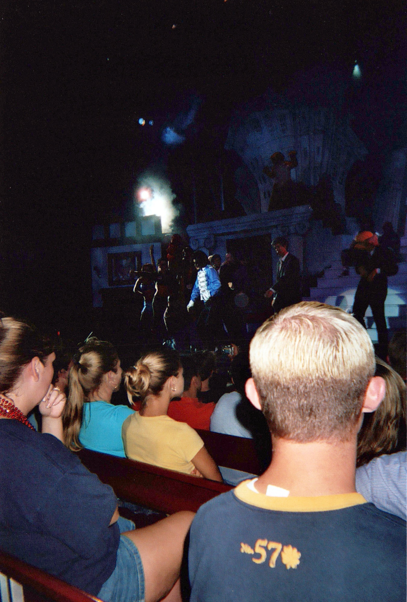 In the foreground crowd watches the stage where Prince, in a blue bandleader jacket, dances in a spotlight. Around prince are the silhouettes of backup dancers.