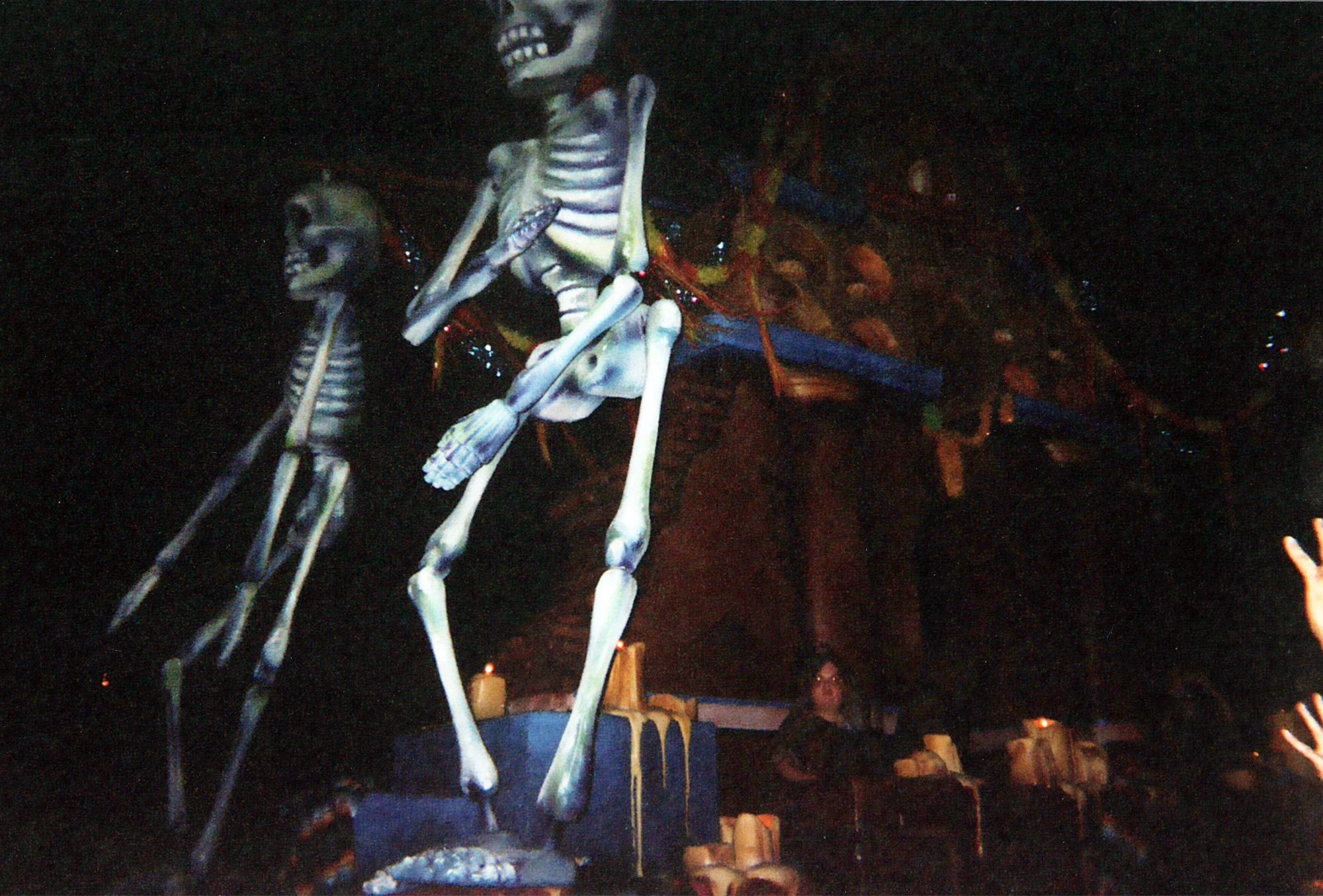 A parade float at night. Two twenty foot skeletons stand in front of of a reddish mausoleum. Dozens of melting candles sit one pillars around its base. Colorful banners hang around a bell tower at the top of the float.