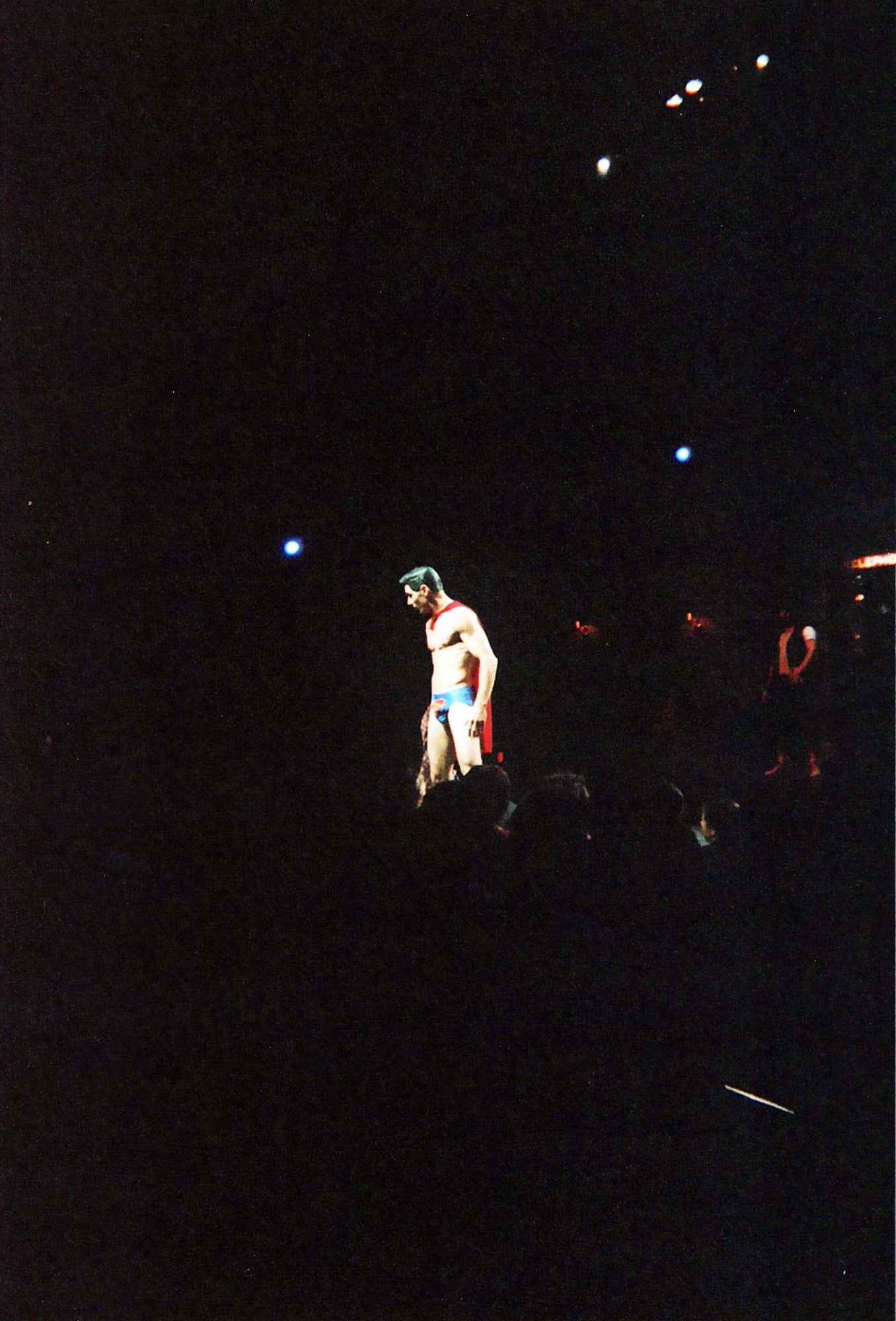 A muscular, disrobed Superman stands alone, lit in the dark. A red cape is tied around his neck. He wears tight, shiny, blue briefs with the Superman “S” symbol across the front.