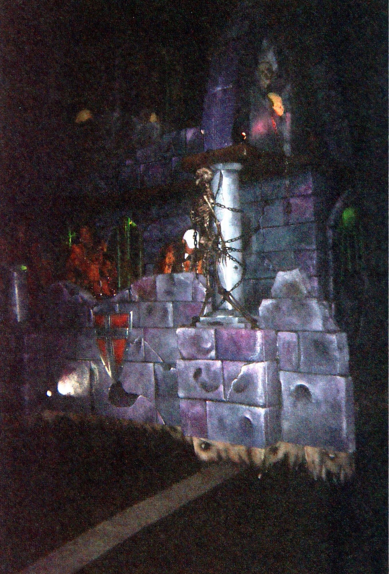 A parade float at night. A purplish grey castle rides along a road. A red shield hangs on a red. Nearby, a gold skeleton is chained to a pillar. Behind the castle wall and pillar, people throw beads. The castle climbs behind them. A barred dungeon glows green on the castle’s other side.