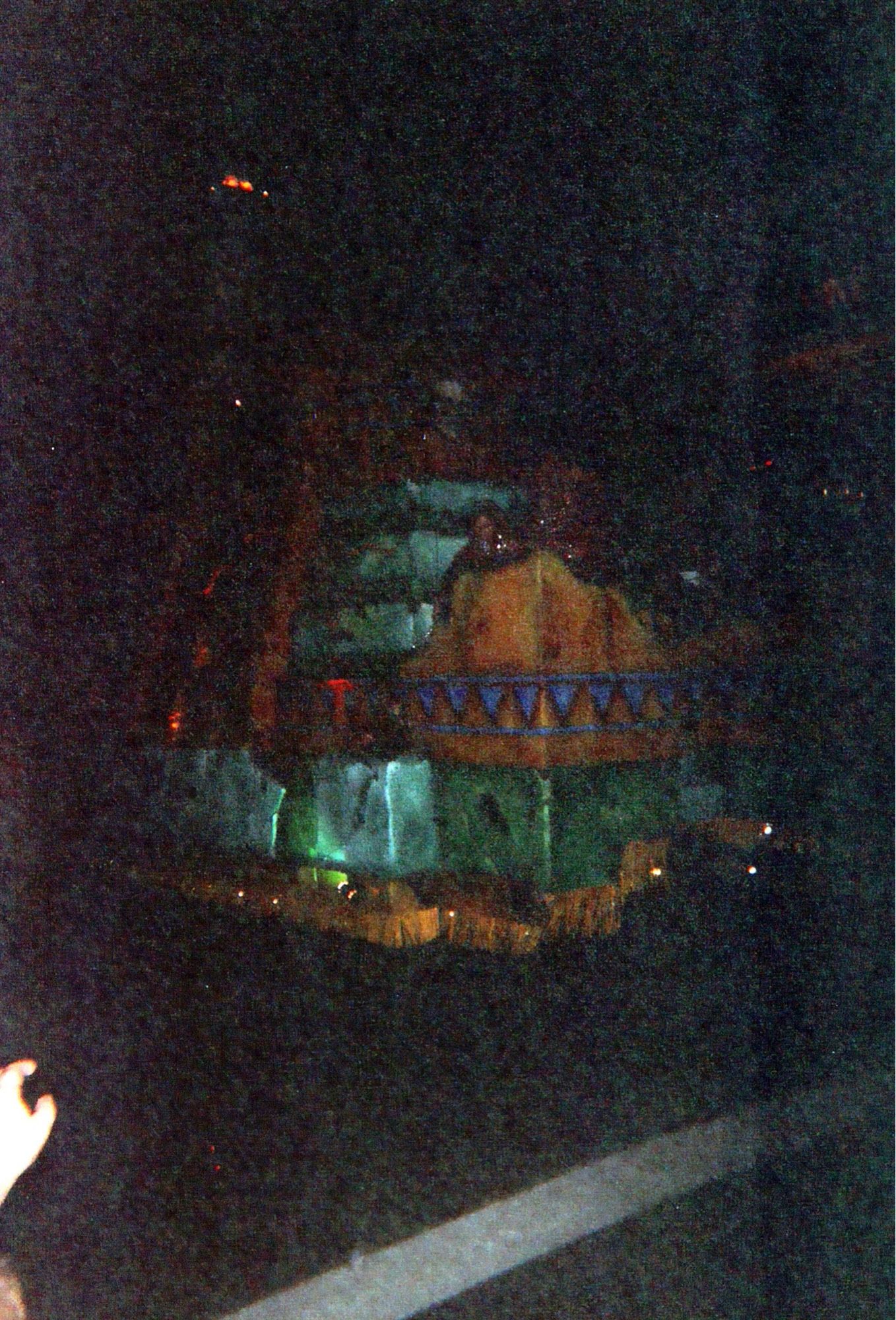 A colorful parade float at night. A green stone temple rides a long a road. At the temple’s side is a broken, yellow wall with a blue, triangular pattern along its base. A woman stands behind the wall.