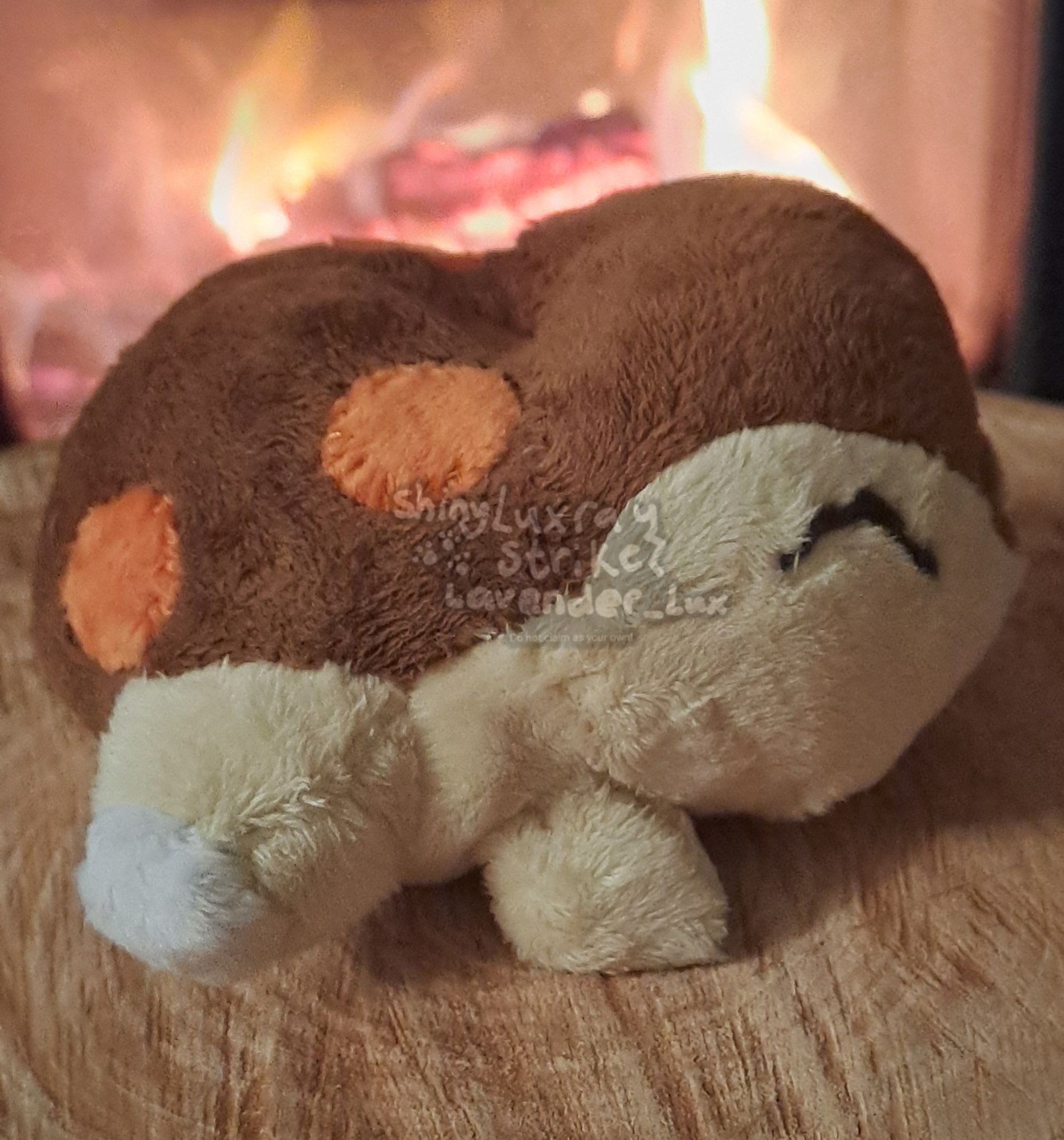 Custom plush of shiny Cyndaquil. Pattern is from a bootleg Cyndaquil pokedoll I seam ripped. 

Cyndaquil, a tan colored pokemon with closed eyes and a brown back with 4 red dots, lies on a piece of wood in front of a fireplace