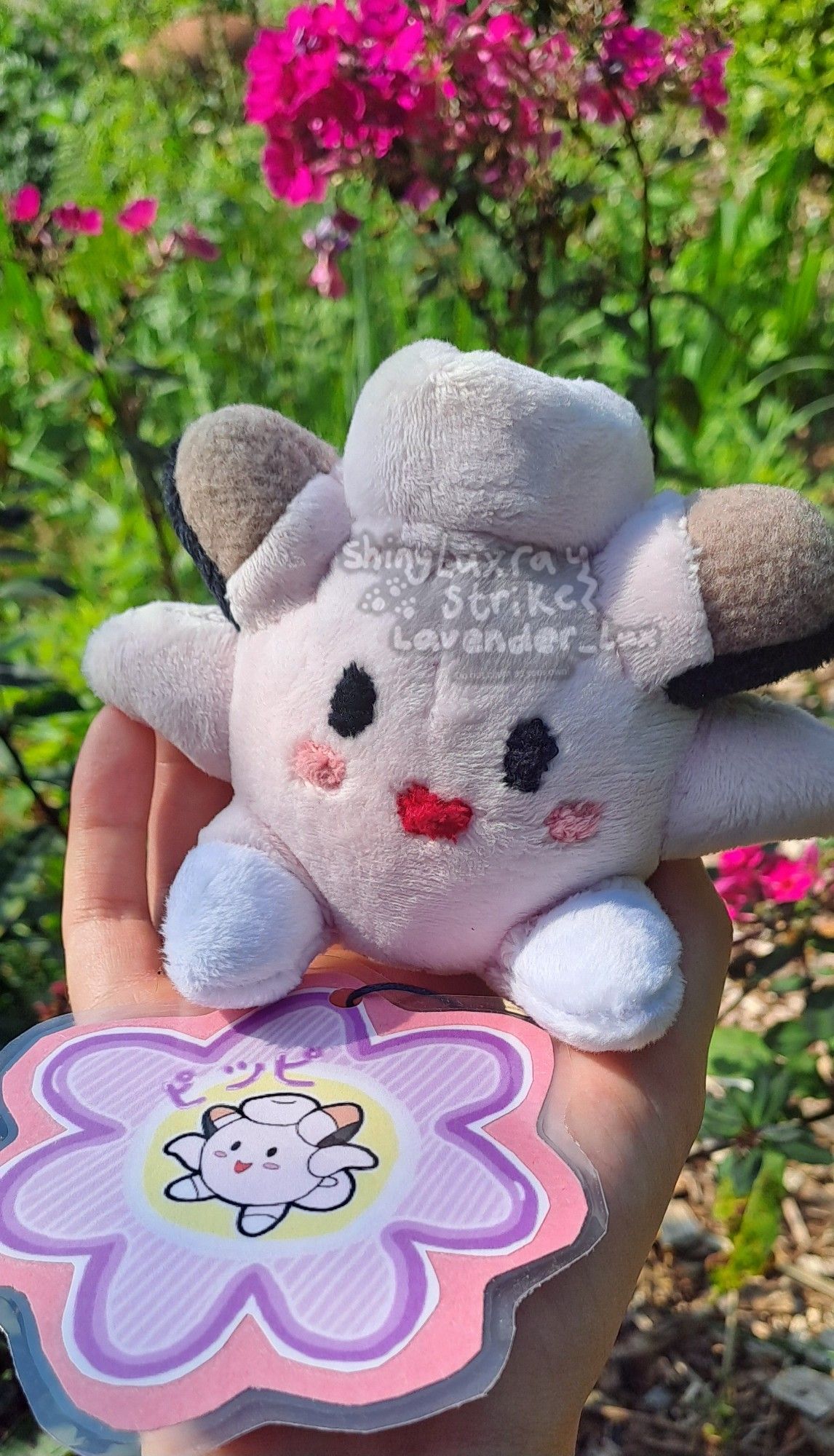 Clefairy mock pokedoll, made to resemble the official pokedoll. Held up againdt some dark pink flowers.