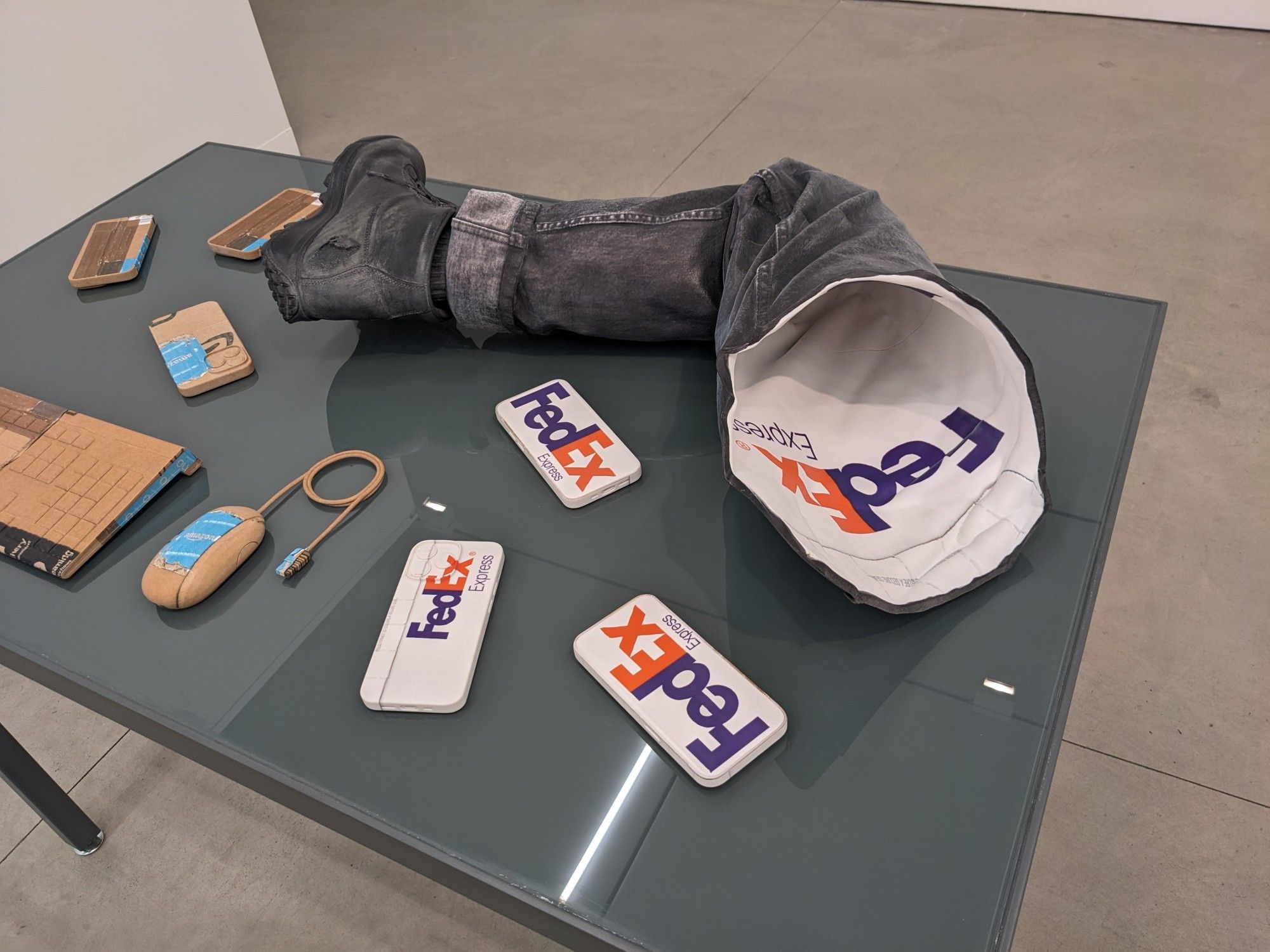 table full of 3d printed full-color Josh Kline sculptures - everyday objects like phones, keyboard, mouse, and a hollow 1:1 scale photoreal leg. But all the objects' textures (in CAD-speak) are FedEx or Amazon package branding