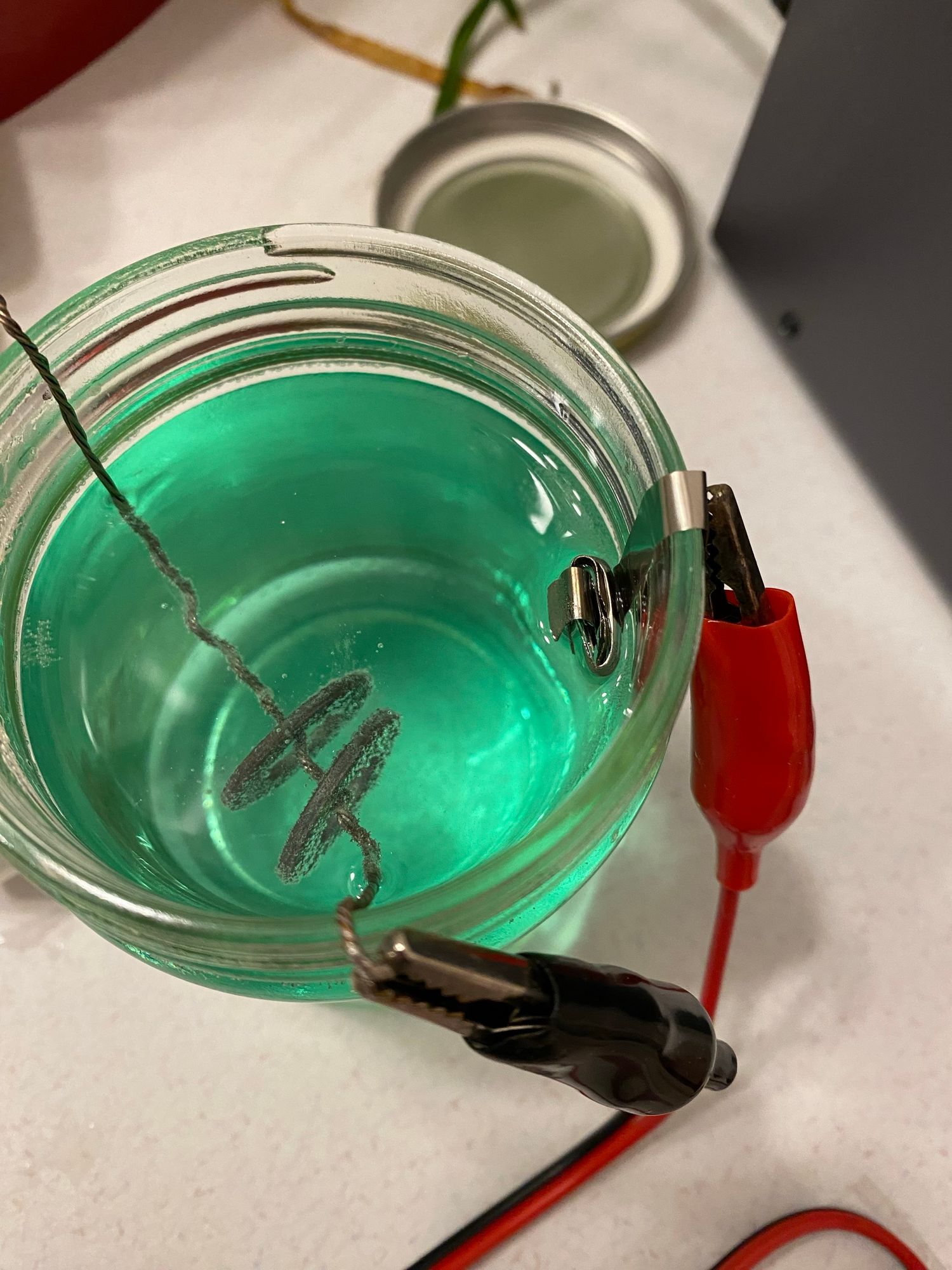 looking down into a glass jar filled with green fluid in which hangs a wire holding two washers. a nickel bundle hangs in the fluid from the jar’s rim. both are connected to wires via alligator clips and the washers are bubbling