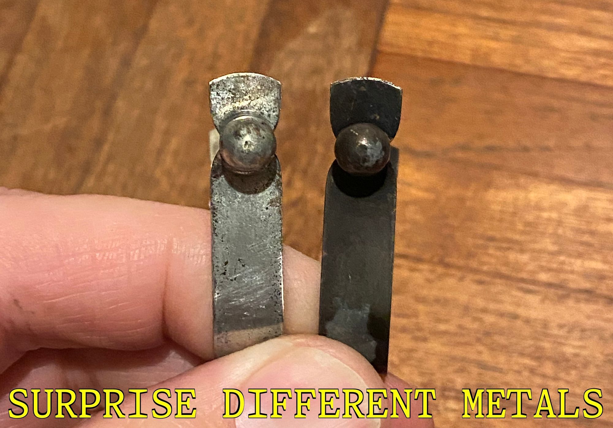 The two identical clips except for clearly being completely different metals as one is nickel grey and the other is now dark black after plating both. The image is presented in the style of a Hand Tool Rescue credit shot, captioned "SURPRISE DIFFERENT METALS" in a close-enough font, in yellow with black outline.