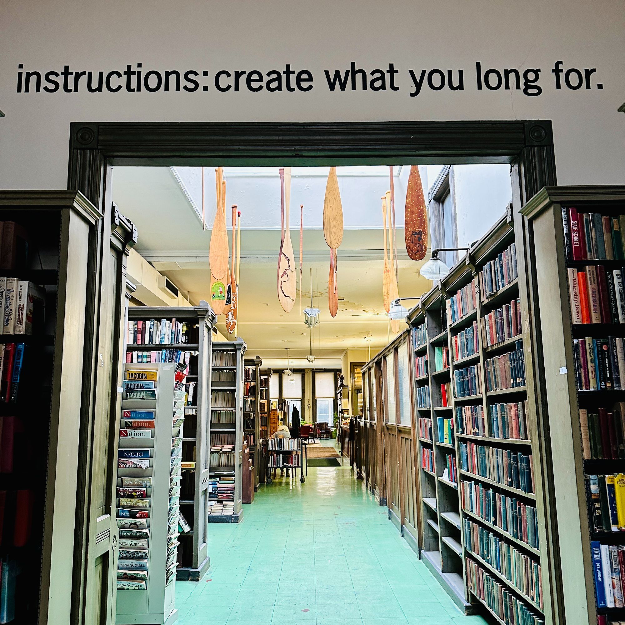 View inside of a library looking through an arch with the words "instructions: create what you long for" written above. Books line the walls and decorated canoe paddles hang from the ceiling.