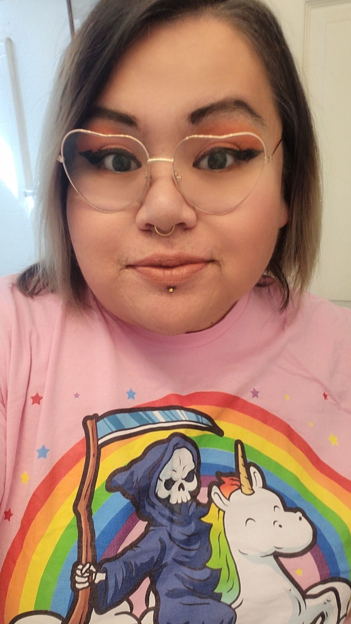 Middle aged woman wearing a pink shirt. Shirt has a cartoon image of death on a unicorn